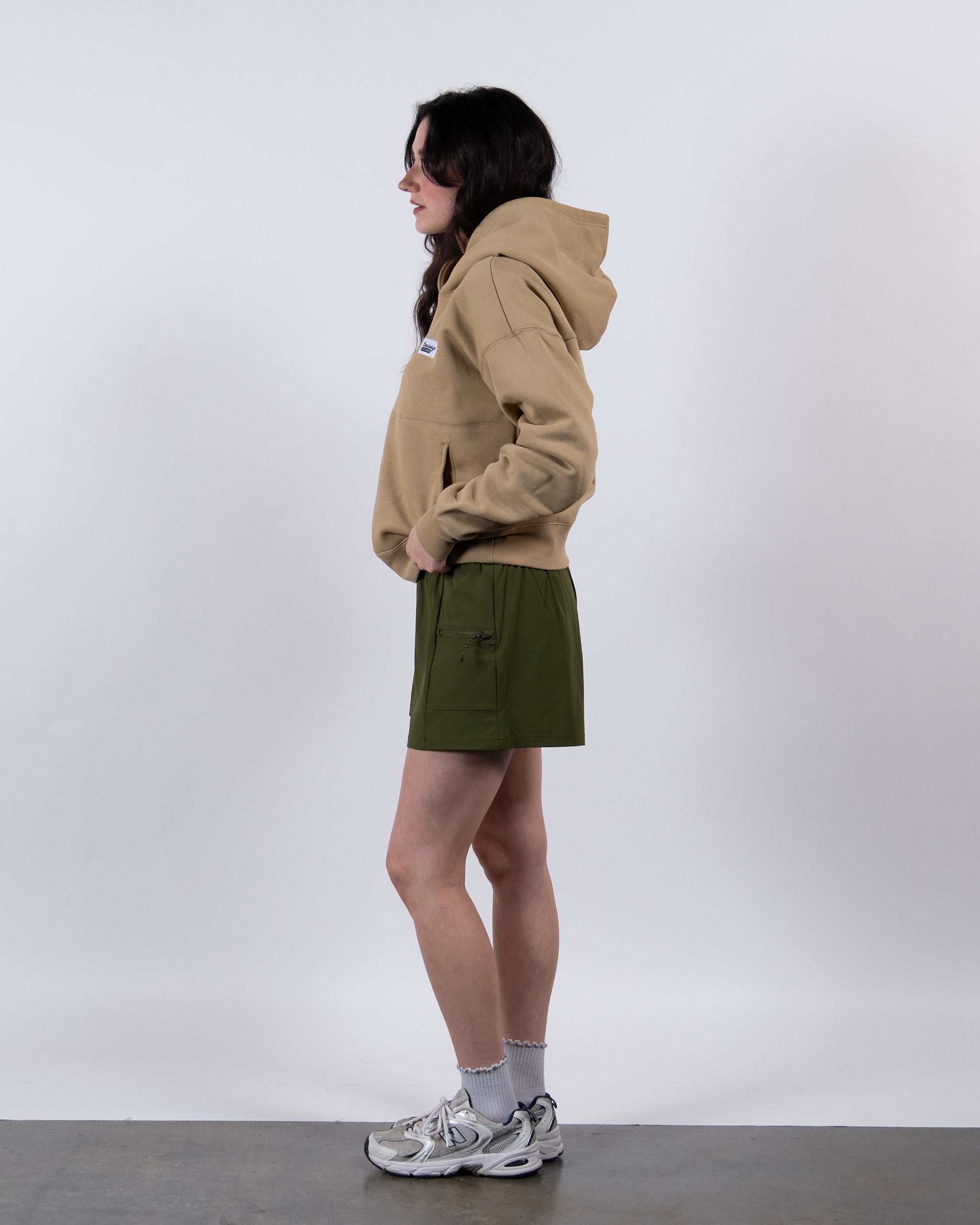 A person stands in profile against a white background, wearing a beardedgoat Women's Overnight™ Hoodie with hands in the pockets, olive green shorts, gray sneakers, white socks, and their dark hair down.