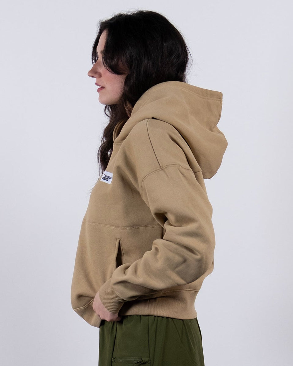 A person stands in profile against a white background, wearing a beardedgoat Women's Overnight™ Hoodie with hands in the pockets, olive green shorts, gray sneakers, white socks, and their dark hair down.