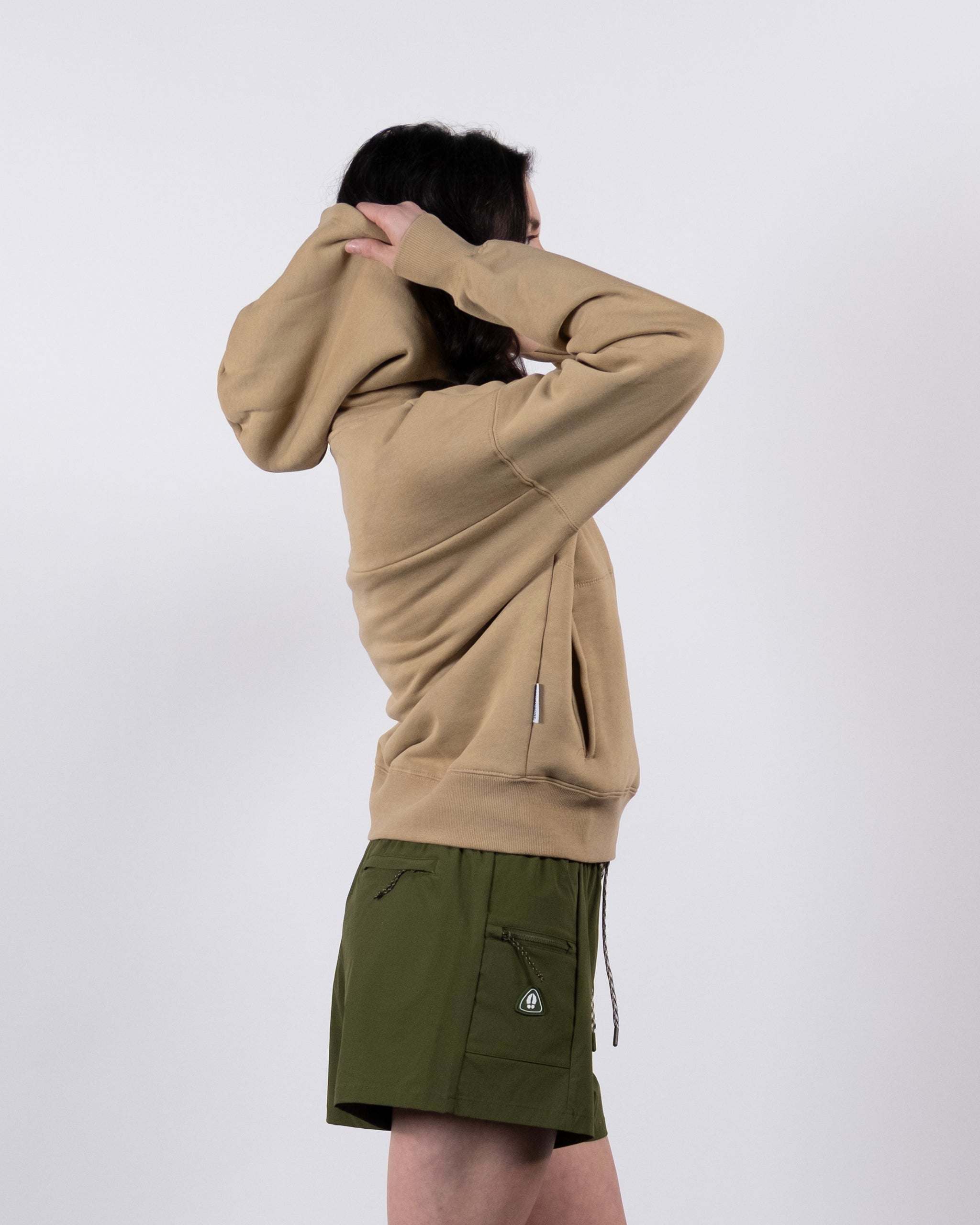 A woman wears a tan beardedgoat Women's Overnight™ Hoodie, crafted from premium cotton fleece, and olive green shorts. She stands in profile against a plain background, raising her hands to adjust the hood.