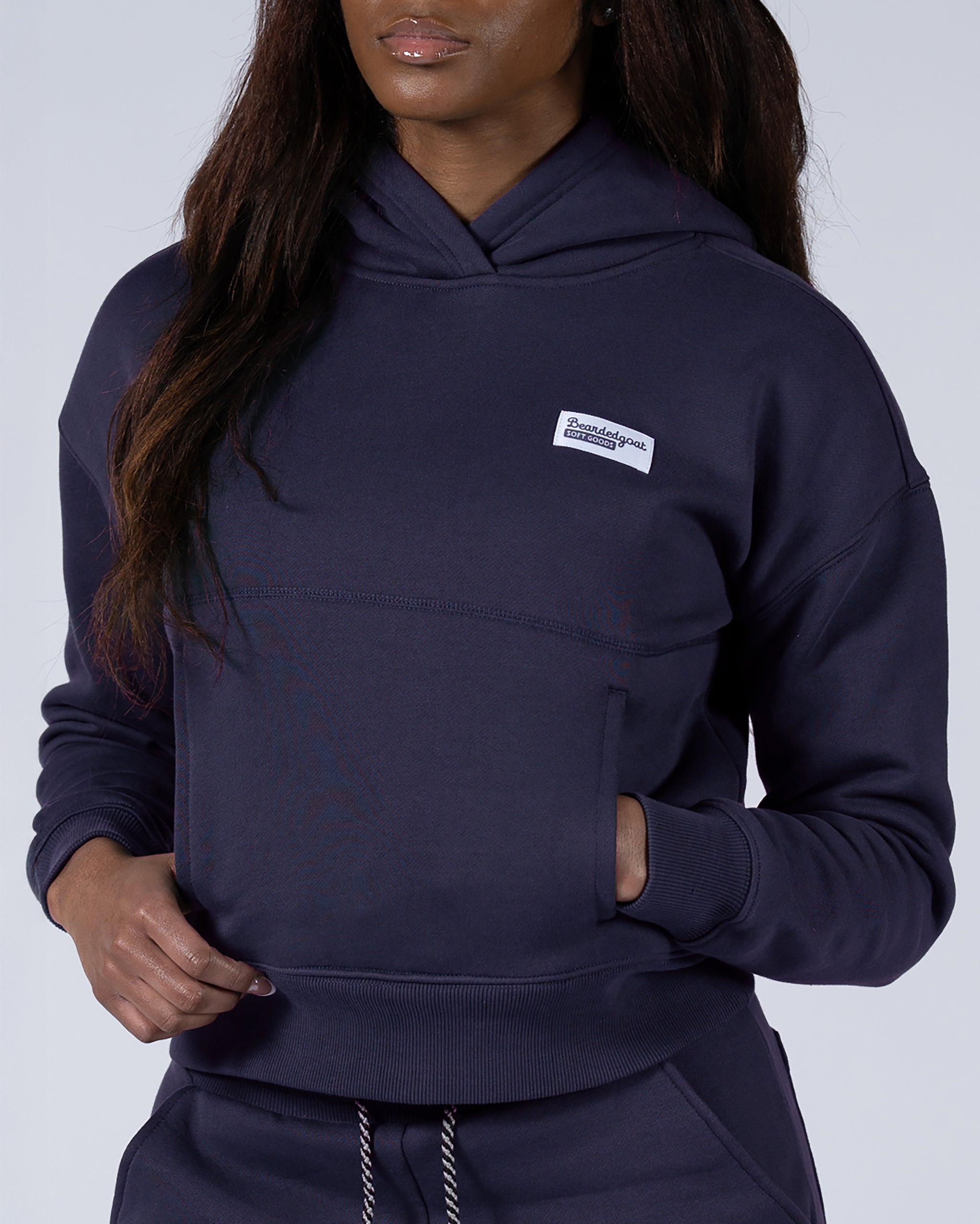 A woman in the beardedgoat Women's Overnight™ Hoodie in Eclipse, featuring premium cotton fleece and a small white chest patch, stands with one hand partly in the pocket. She has long, straight hair and wears matching joggers.