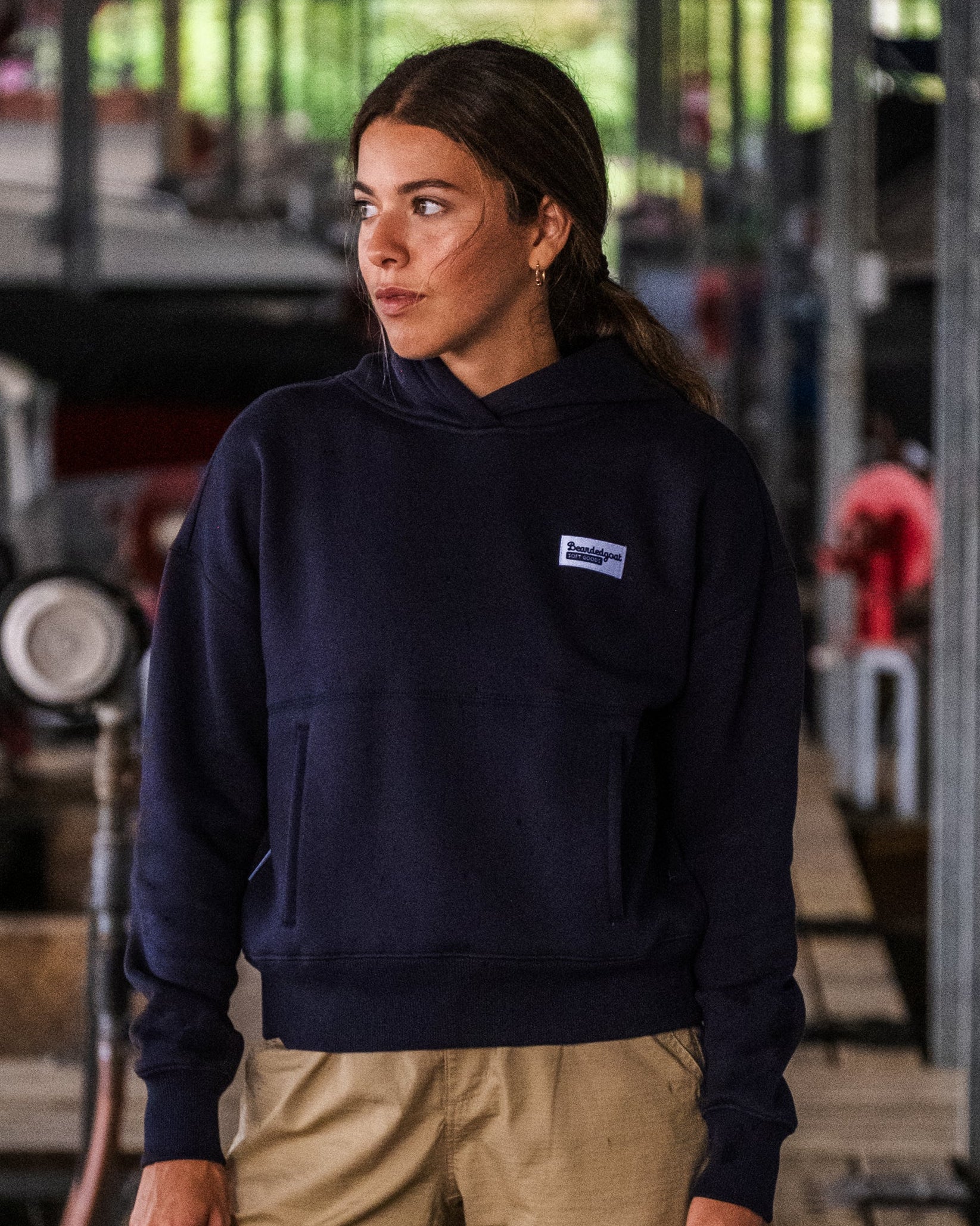 A woman wearing the beardedgoat Women's Overnight™ Hoodie and beige pants stands on a wooden dock in an industrial setting, gazing thoughtfully to the side.