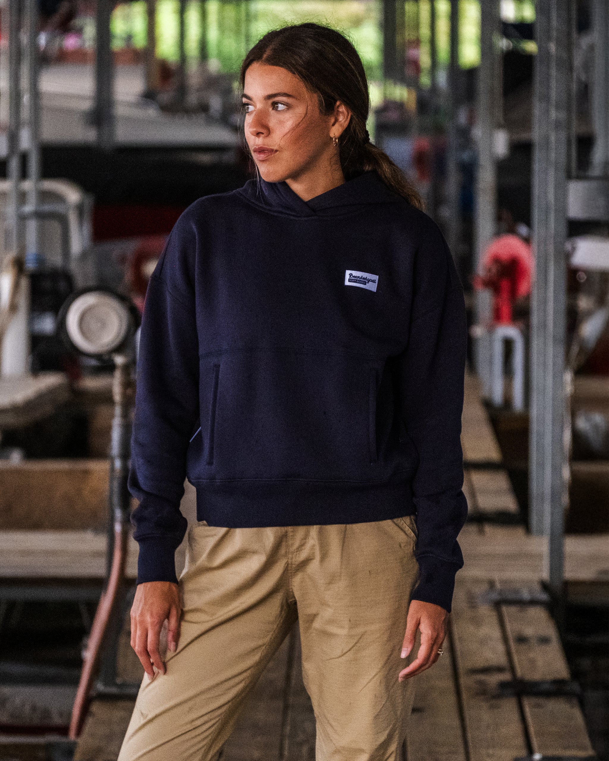 A woman wears the beardedgoat Women's Overnight Hoodie and beige pants, standing thoughtfully on a wooden dock with industrial machinery and metal structures in the background.
