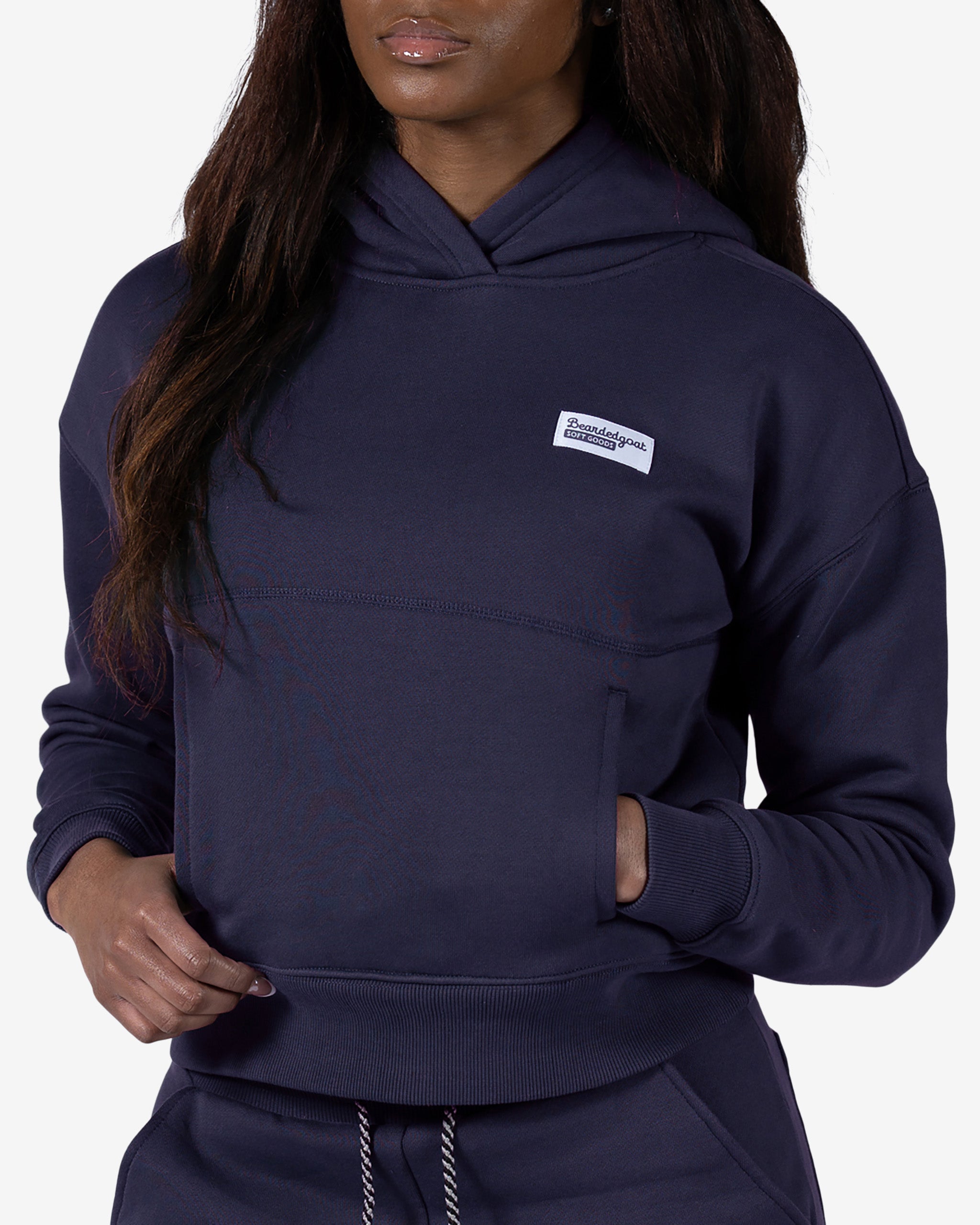 A woman wears the beardedgoat Women's Overnight™ Hoodie, a relaxed fit premium cotton fleece with a small rectangular white chest patch and front pocket, posing with one hand near her waist.