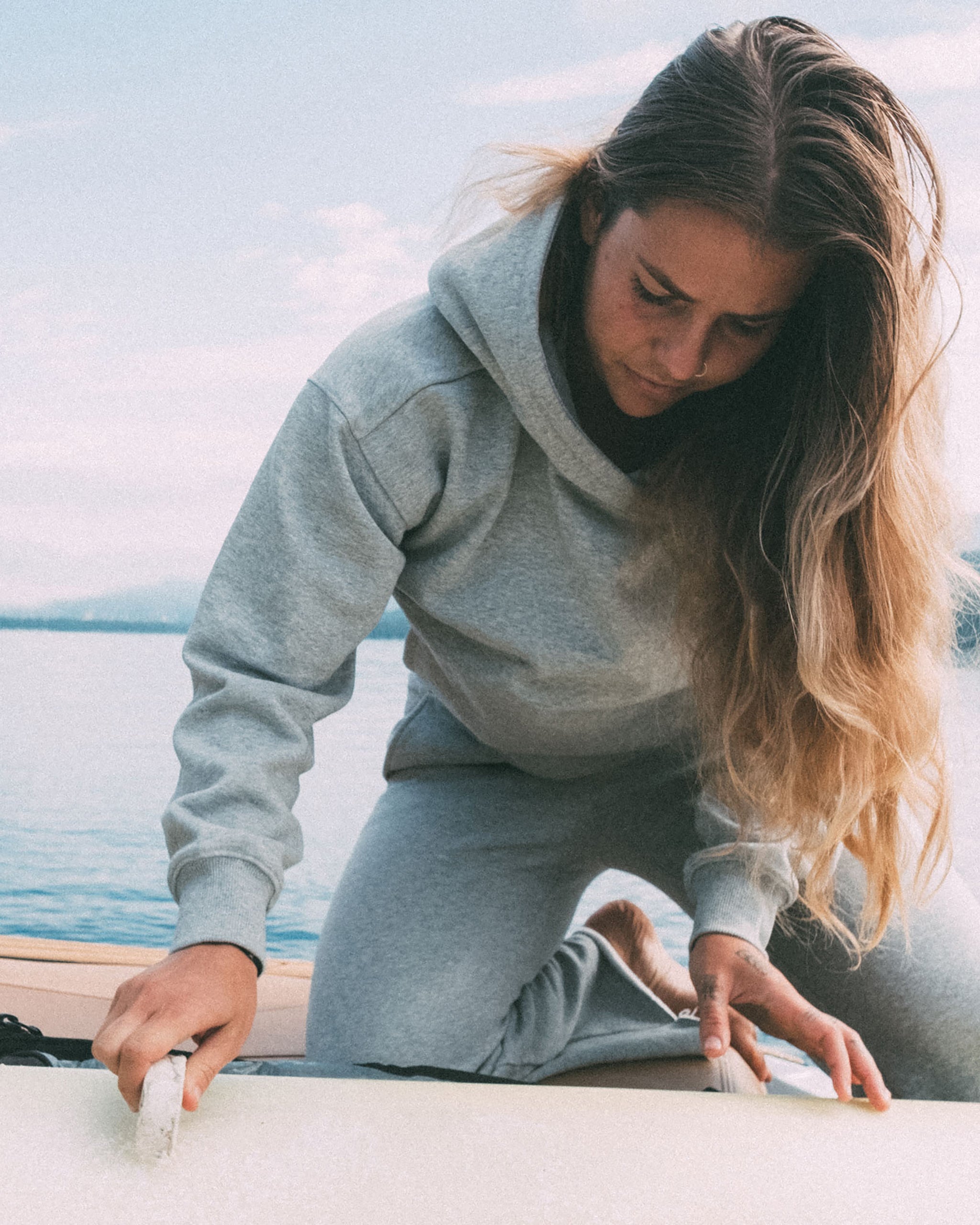 Wearing the beardedgoat Women's Overnight Hoodie, she kneels on a paddleboard by the water, waxing its surface with her long hair falling to one side in a calm outdoor setting.