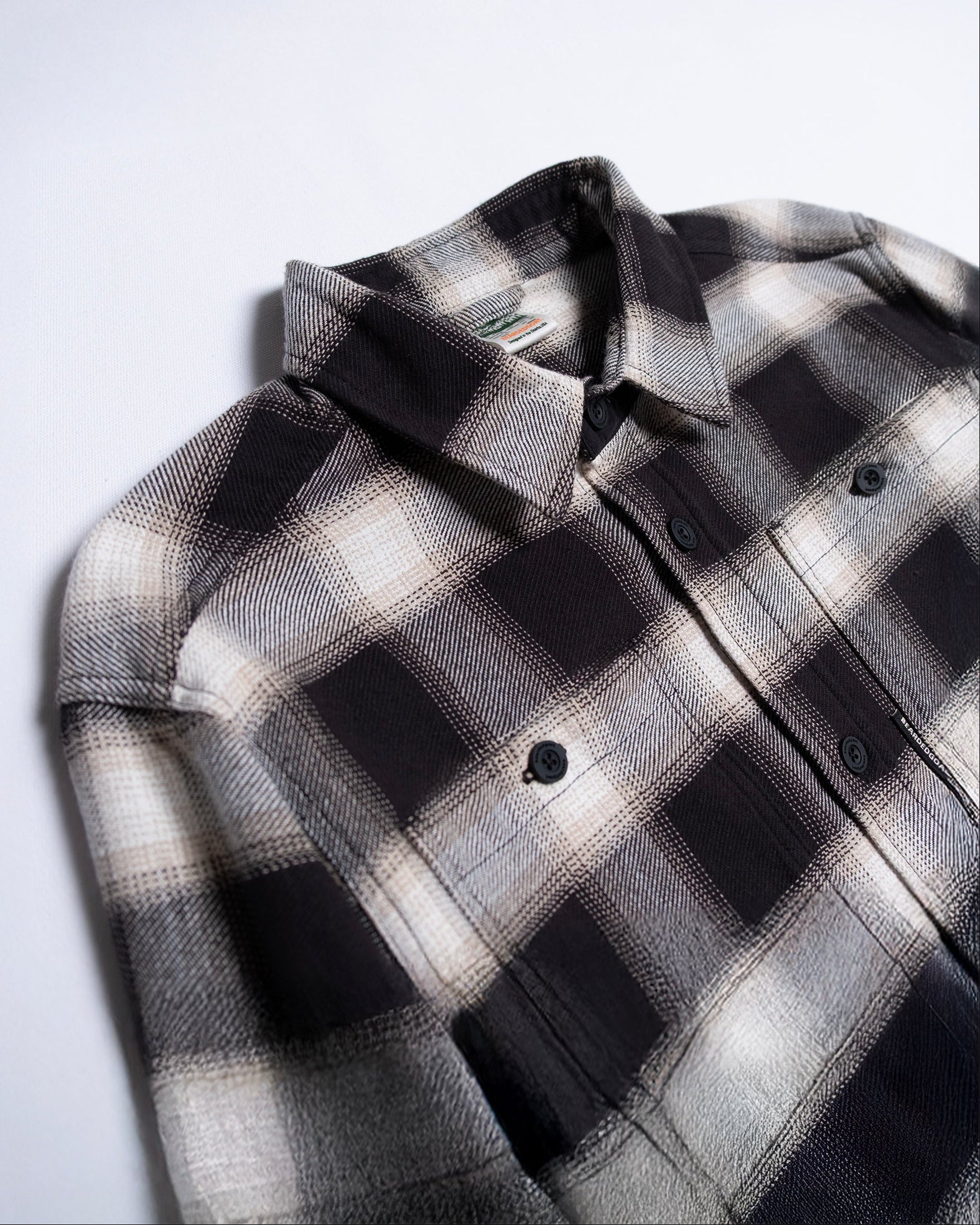 A close-up of the beardedgoat NEW--Ozark Utility Flannel™ in Black, a durable cotton long-sleeve button-up shirt with a black plaid pattern and two buttoned chest pockets, laid flat on a white surface.
