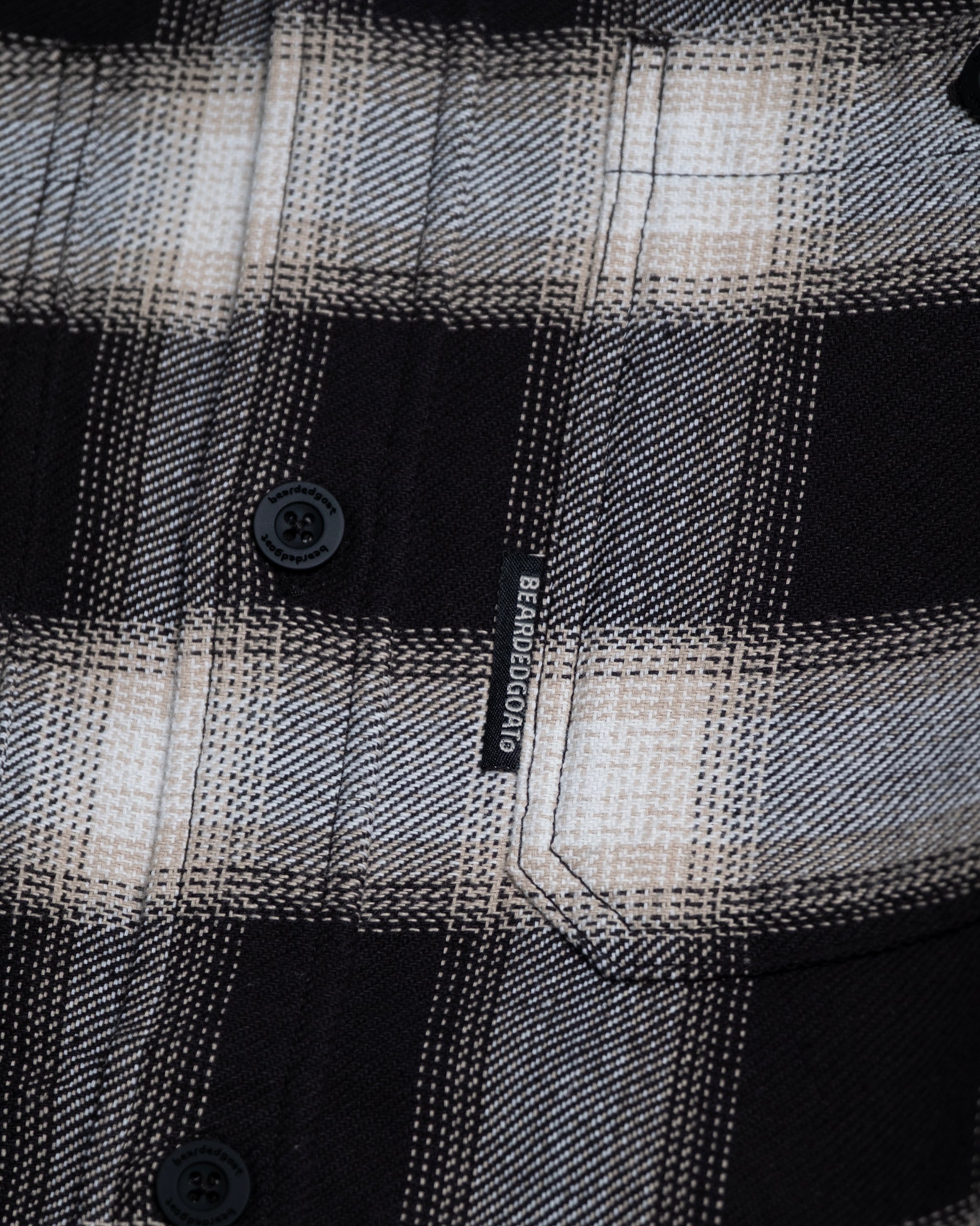 Close-up of the NEW--Ozark Utility Flannel™ by beardedgoat, featuring a black and white plaid pattern, black buttons, and pocket. A small black tag with white text is sewn into the pocket seam of this durable work shirt.