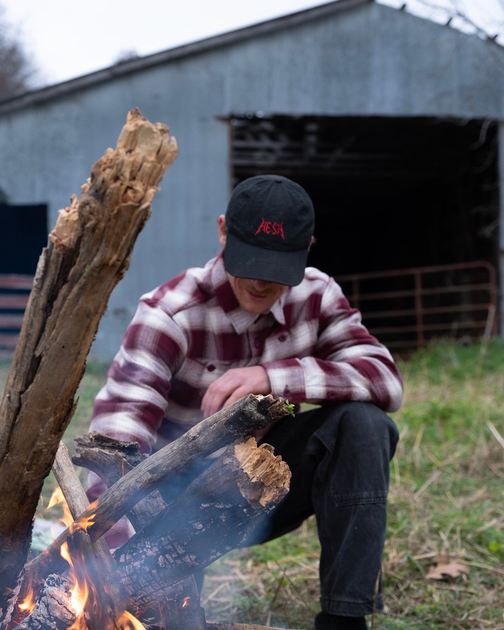 Wearing the beardedgoat NEW--Ozark Utility Flannel™ and a black cap, a person crouches by a campfire, tending to burning logs with a weathered metal barn in the grassy background.