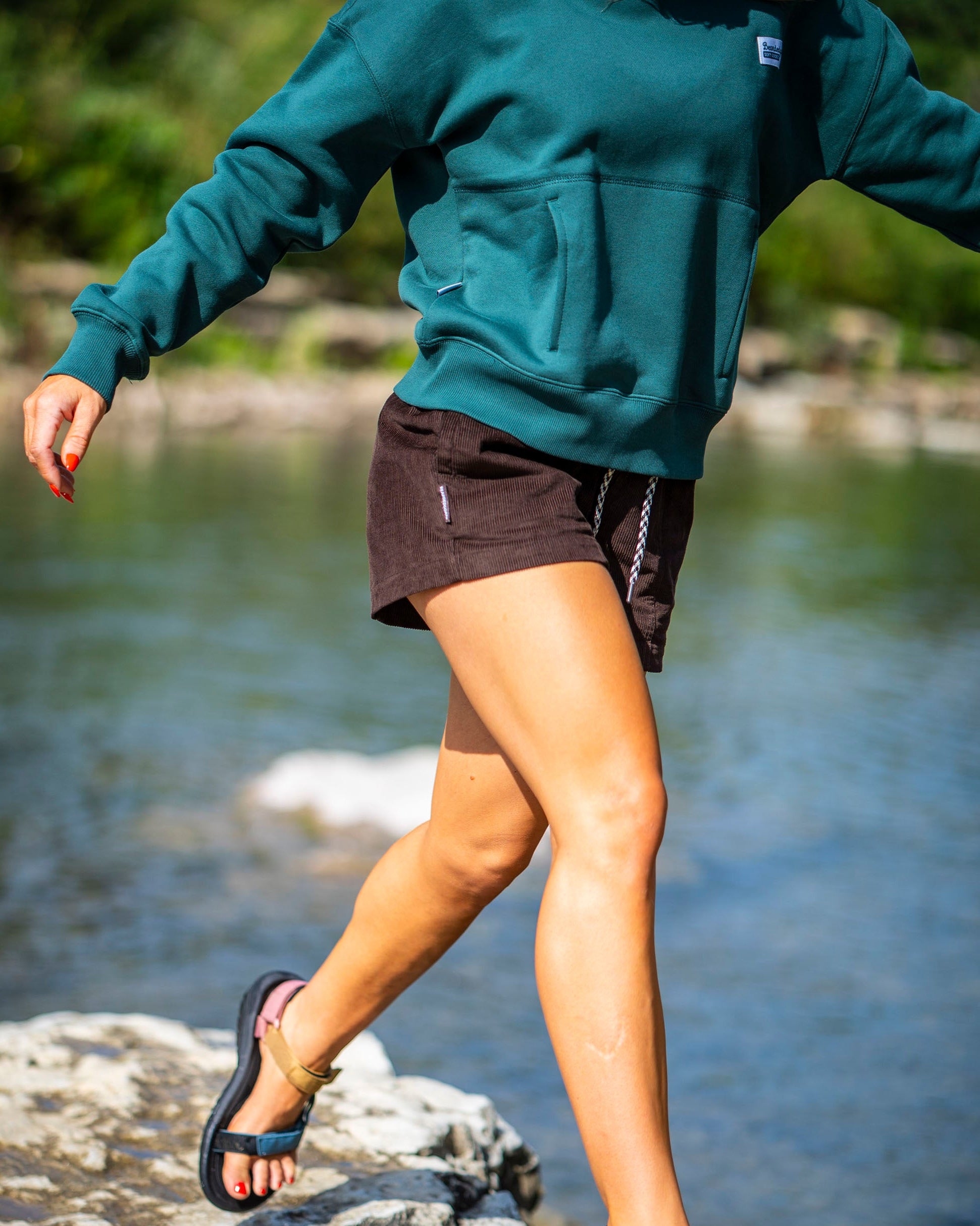Wearing BEARDEDGOAT Women's Cord Shorts, a person in a green hoodie and sandals steps over stream rocks, sunlight highlighting their legs and the scenic outdoors.