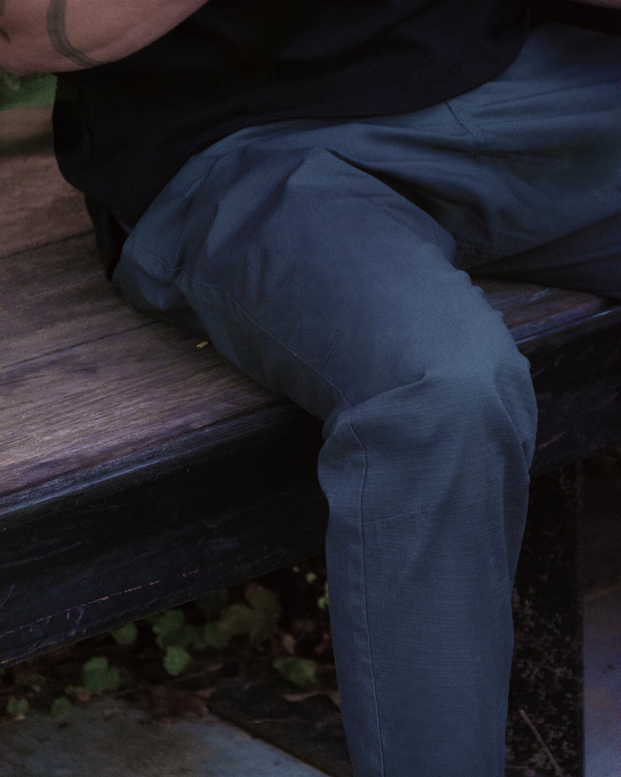 A person wearing BEARDEDGOAT’s NEW Men's Canopy™ Pant made from durable stretch cotton and a black shirt sits on a wooden bench outside, with only their lower torso and legs visible.