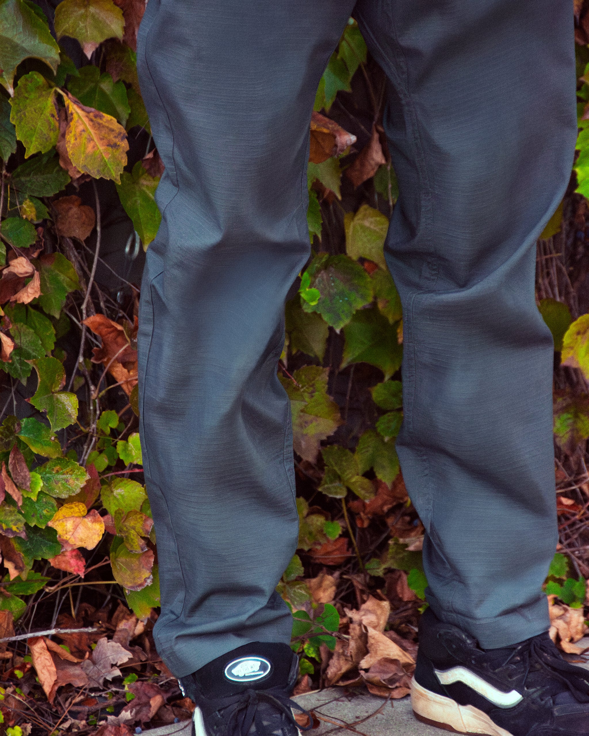 A person wearing BEARDEDGOAT's NEW - Men's Canopy™ Pant in gray and black sneakers stands on a sidewalk with colorful autumn leaves and green vines in the background.