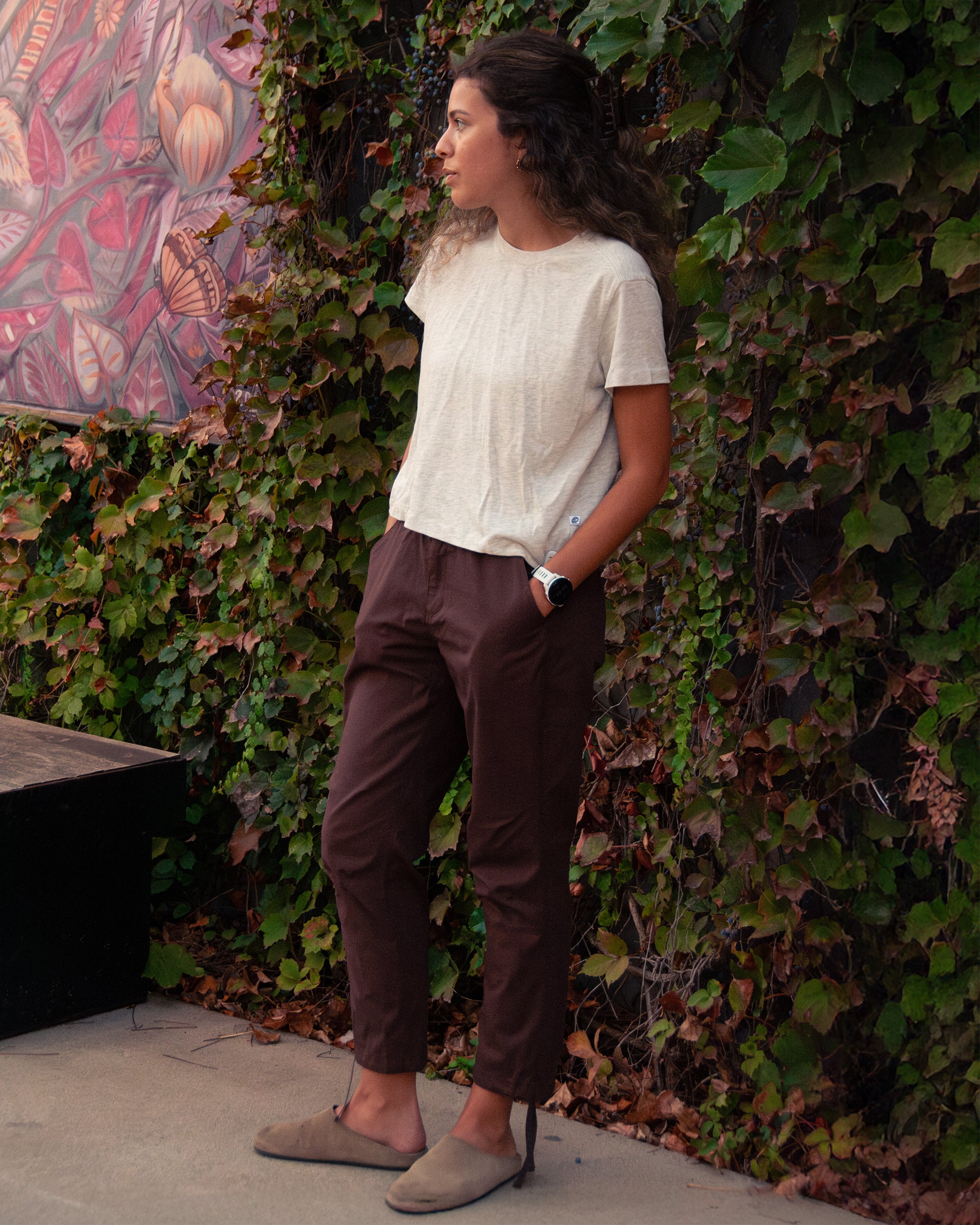 A woman with wavy hair stands outdoors against green ivy, wearing BEARDEDGOAT Women's Canopy™ Pant in stretch cotton ripstop, a white tee, tan shoes, and a watch. She looks to the side with hands in her pockets.