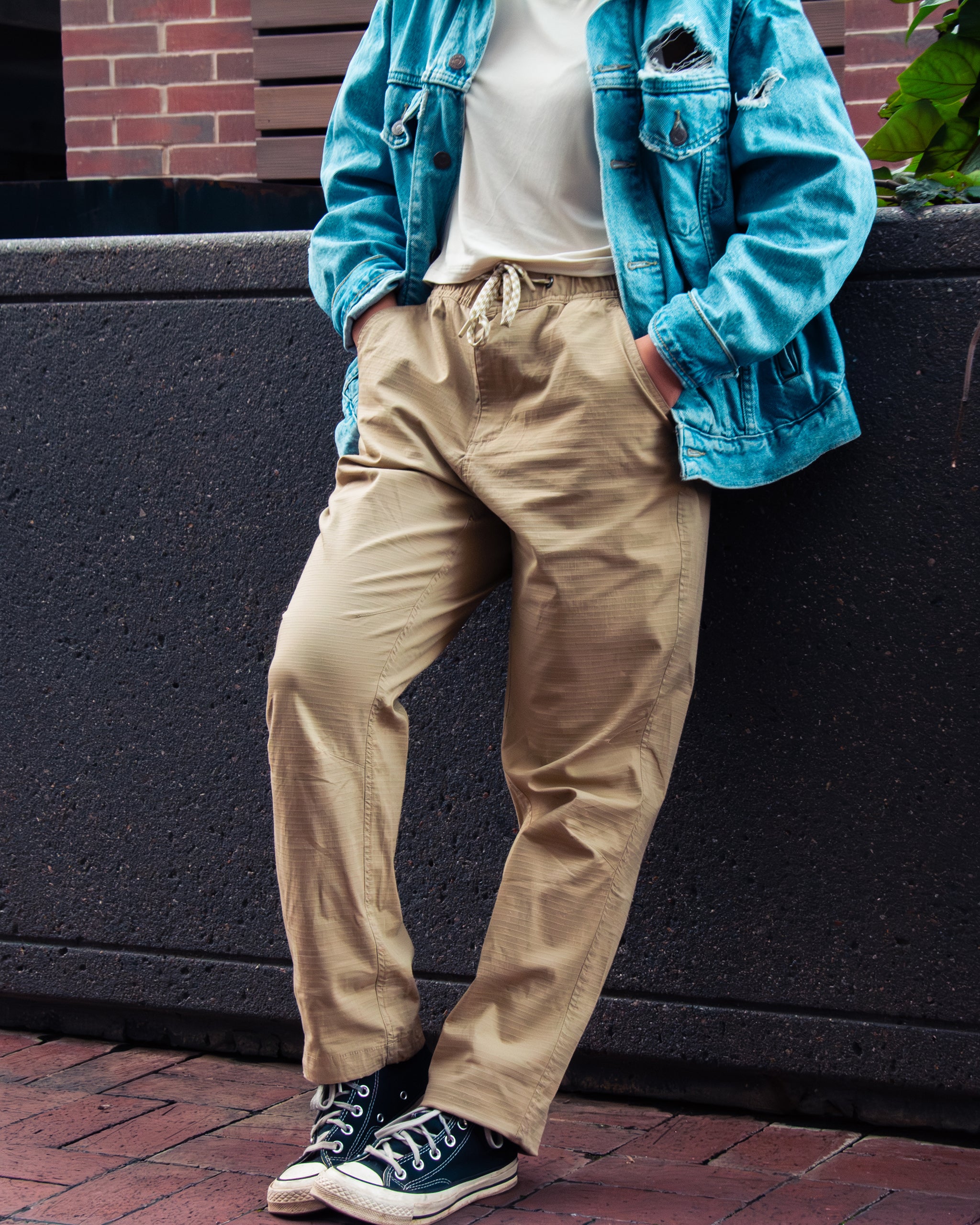 A woman stands with hands in pockets, wearing BEARDEDGOAT's Women's Canopy™ Pant in beige stretch cotton ripstop, a white shirt, blue denim jacket, and black Converse sneakers, leaning against a dark stone wall.