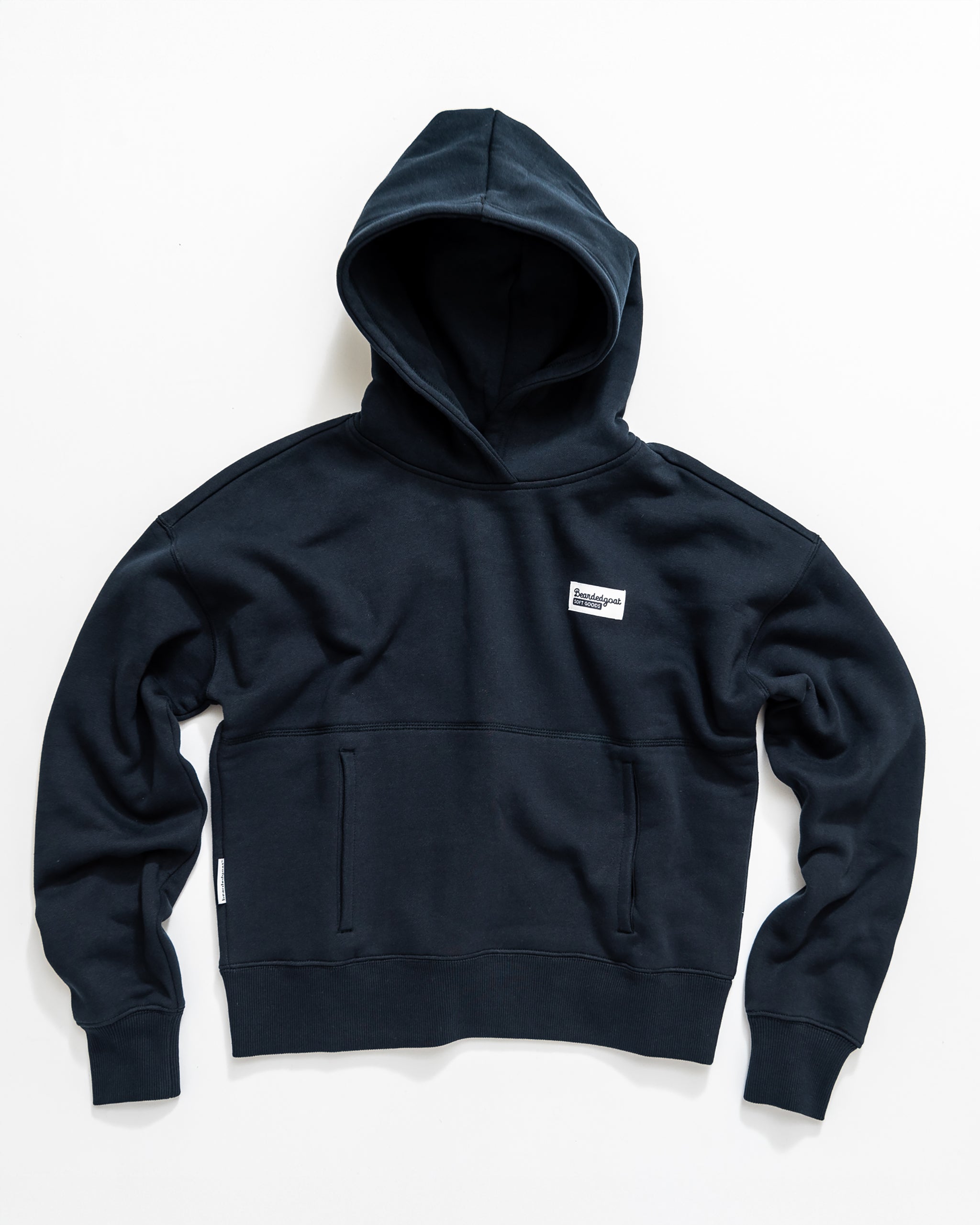 The beardedgoat Women's Overnight Hoodie is a navy blue heavyweight cotton fleece sweatshirt with a front kangaroo pocket and a small white rectangular chest logo, shown laid flat on a white background.
