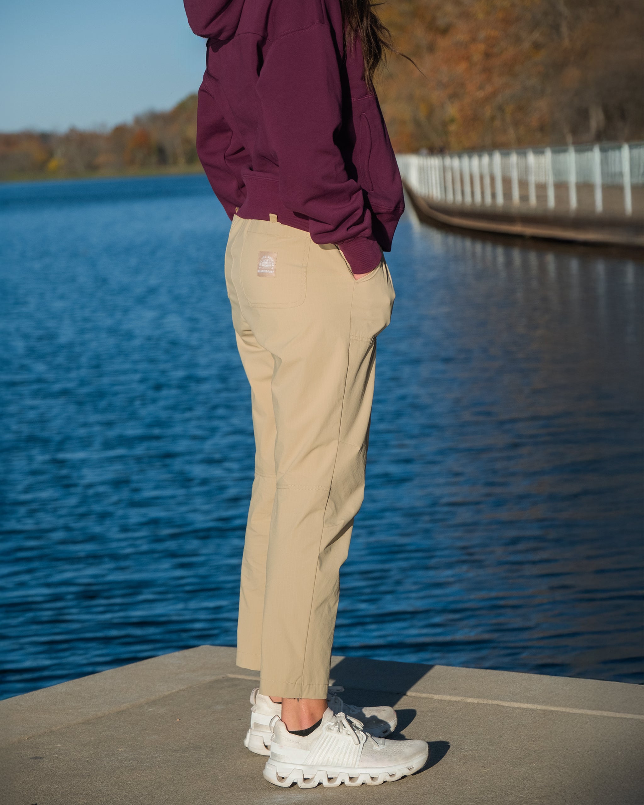 A person stands on a pier by the water, wearing a maroon hoodie, BEARDEDGOAT NEW — Summit™ Pant, and white sneakers. Autumn trees and a curved shoreline railing create a scenic background.