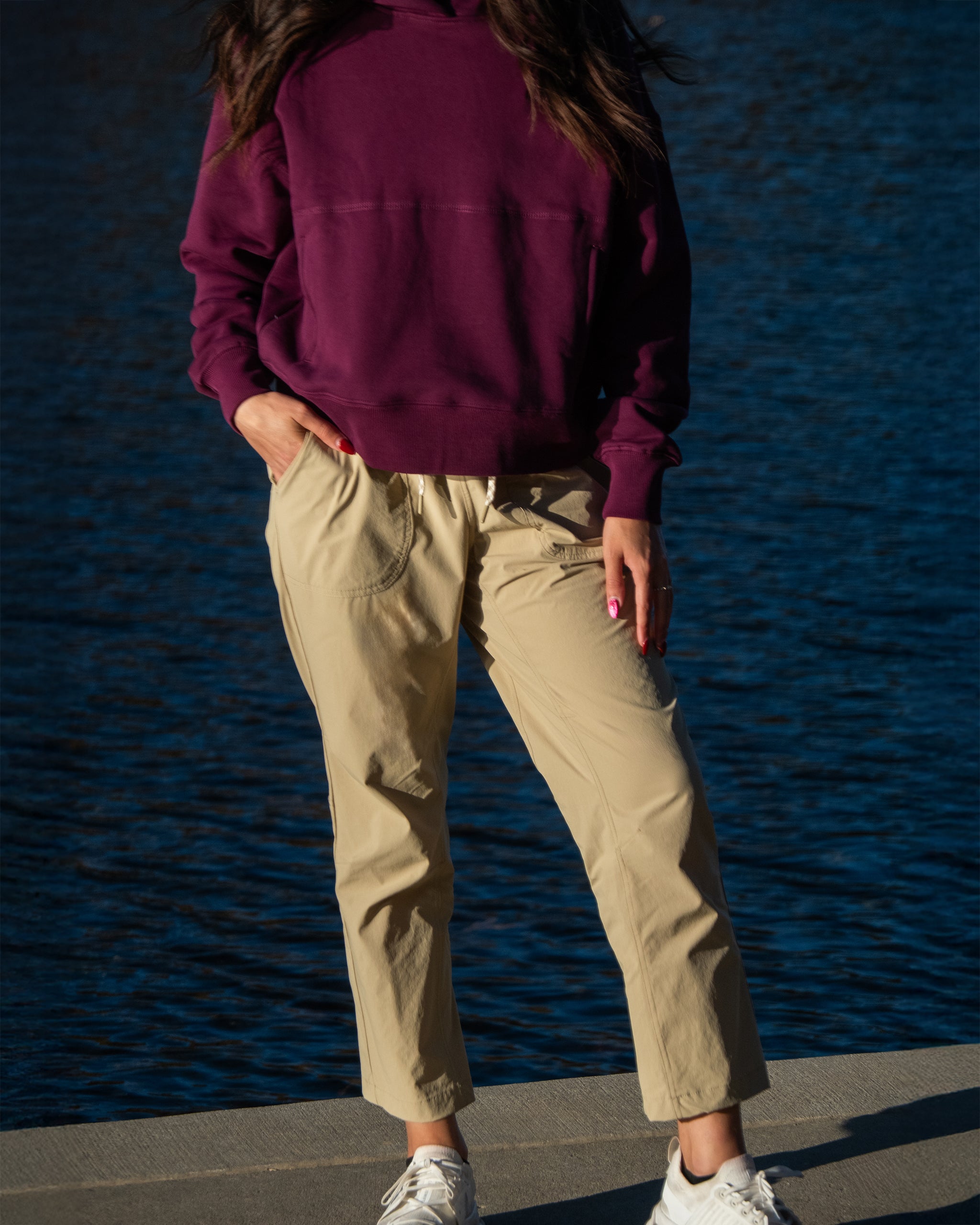 A person stands outdoors by water, wearing a maroon hoodie, white sneakers, and BEARDEDGOAT's NEW — Summit™ Pant. Their hands are in their pockets and their face is not visible.