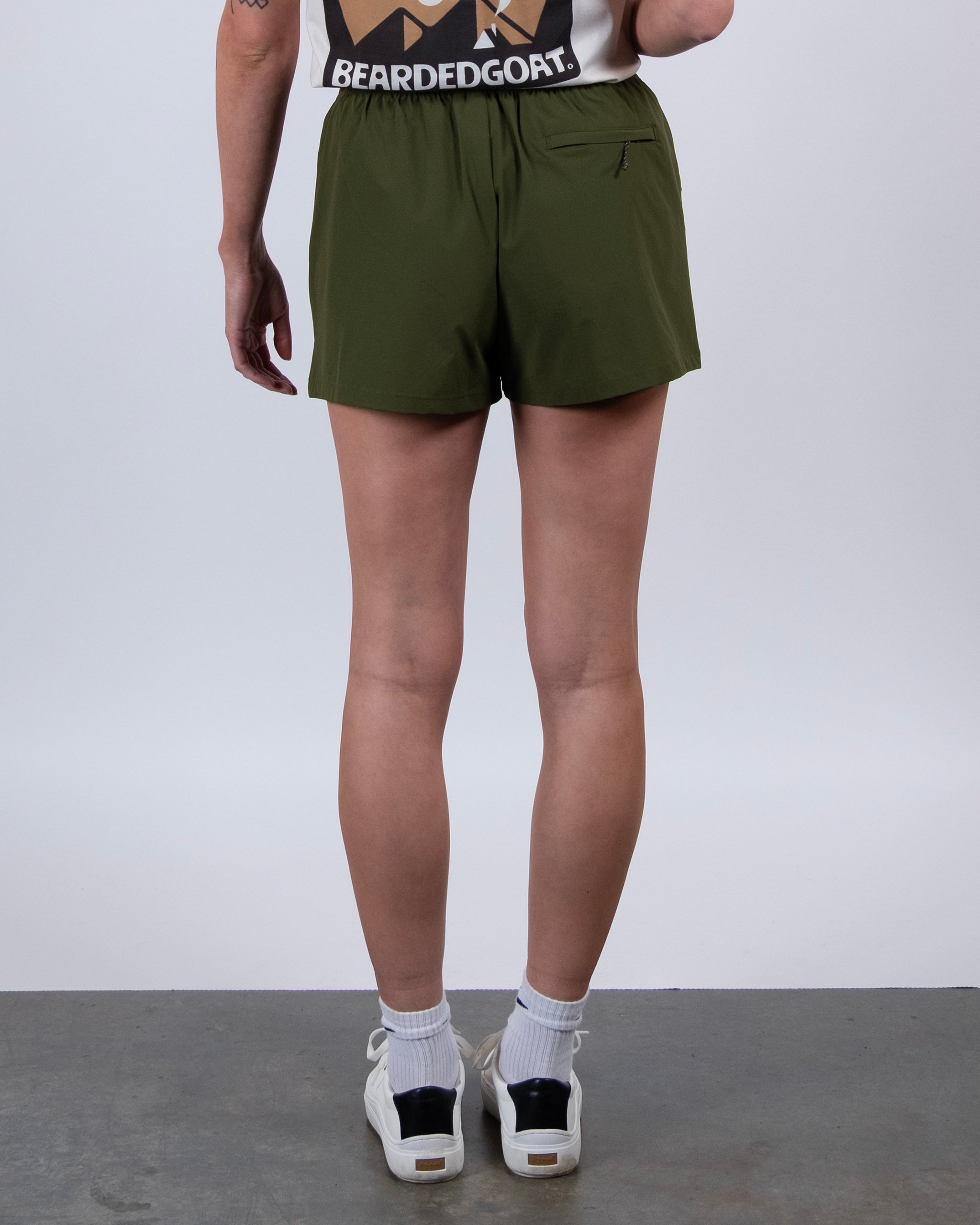 A person stands on a concrete floor facing away, wearing BEARDEDGOAT's olive green Summit Short made from lightweight ripstop fabric, a sleeveless BEARDEDGOAT shirt, white ankle socks, and white sneakers.