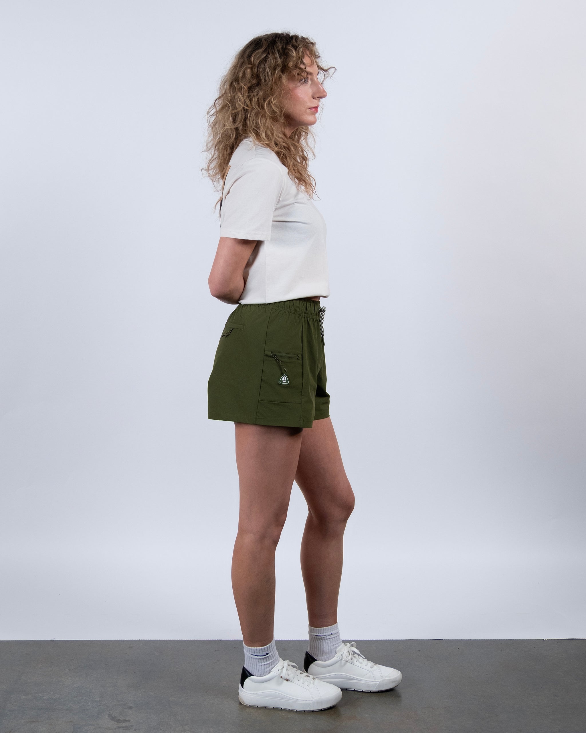A person with curly hair stands in profile against a plain light background, wearing a short-sleeved white shirt, BEARDEDGOAT Summit Short in green lightweight ripstop fabric, white socks, and white sneakers, arms behind their back.