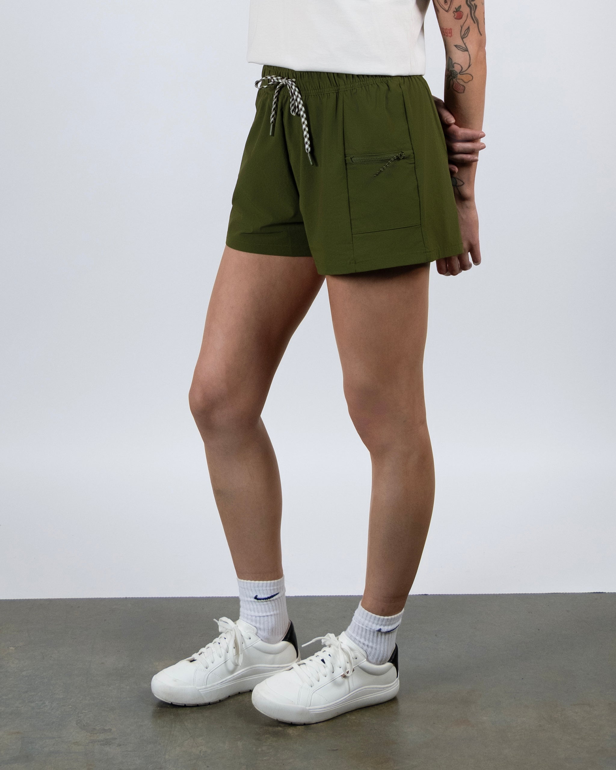 A person stands on a gray floor wearing BEARDEDGOAT Summit Shorts in olive green, crafted from lightweight ripstop with a front pocket and white drawstring. Only the lower half and one tattooed arm are visible, paired with white socks and sneakers.