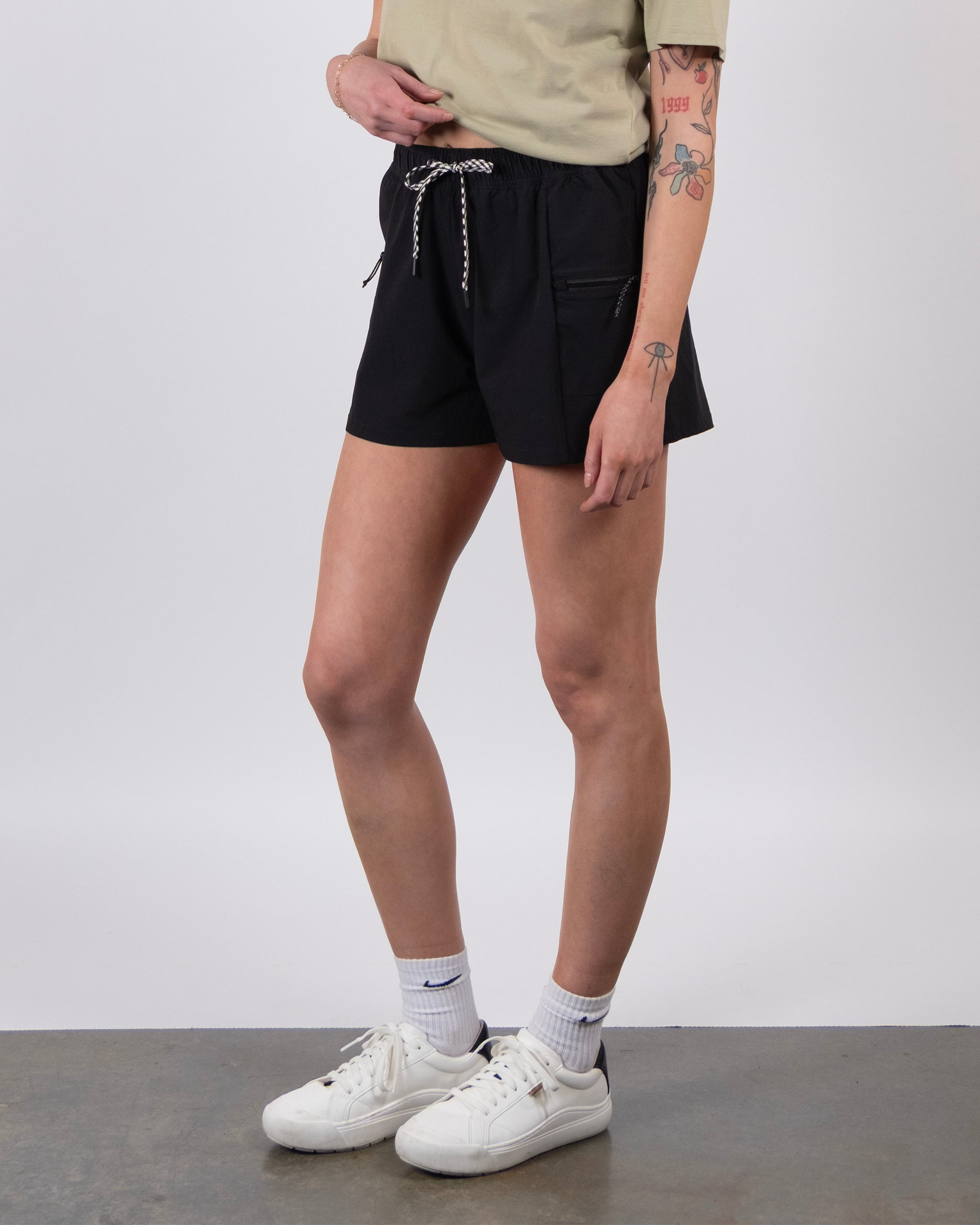 A person stands indoors on a gray concrete floor wearing BEARDEDGOAT's black Summit Short (lightweight ripstop fabric), a beige t-shirt, white socks, and white sneakers, with visible tattoos on their left arm and hand.