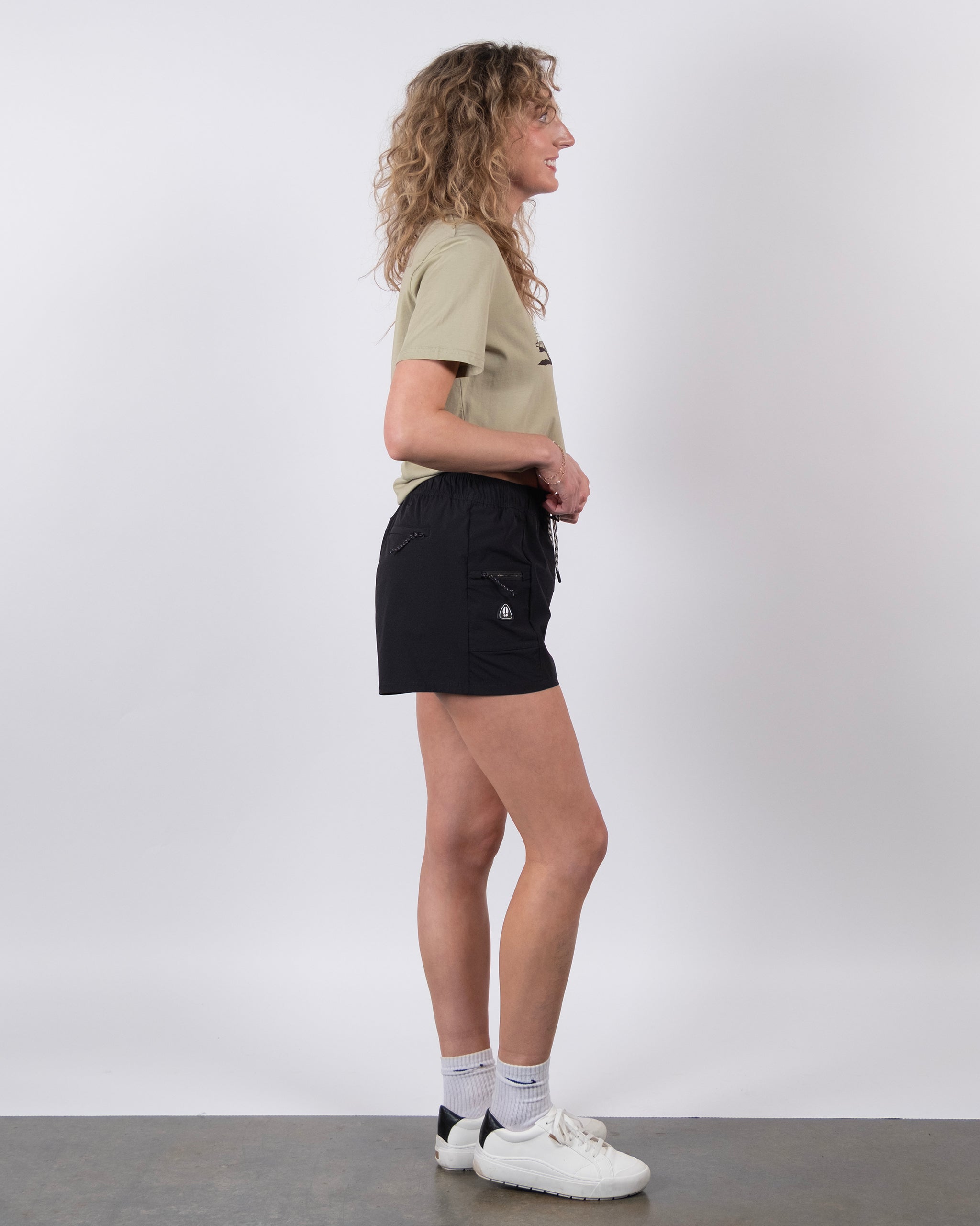 A woman with curly hair stands in profile against a white background, wearing a beige t-shirt, BEARDEDGOAT Summit Shorts made from lightweight ripstop fabric, white socks, and white sneakers.