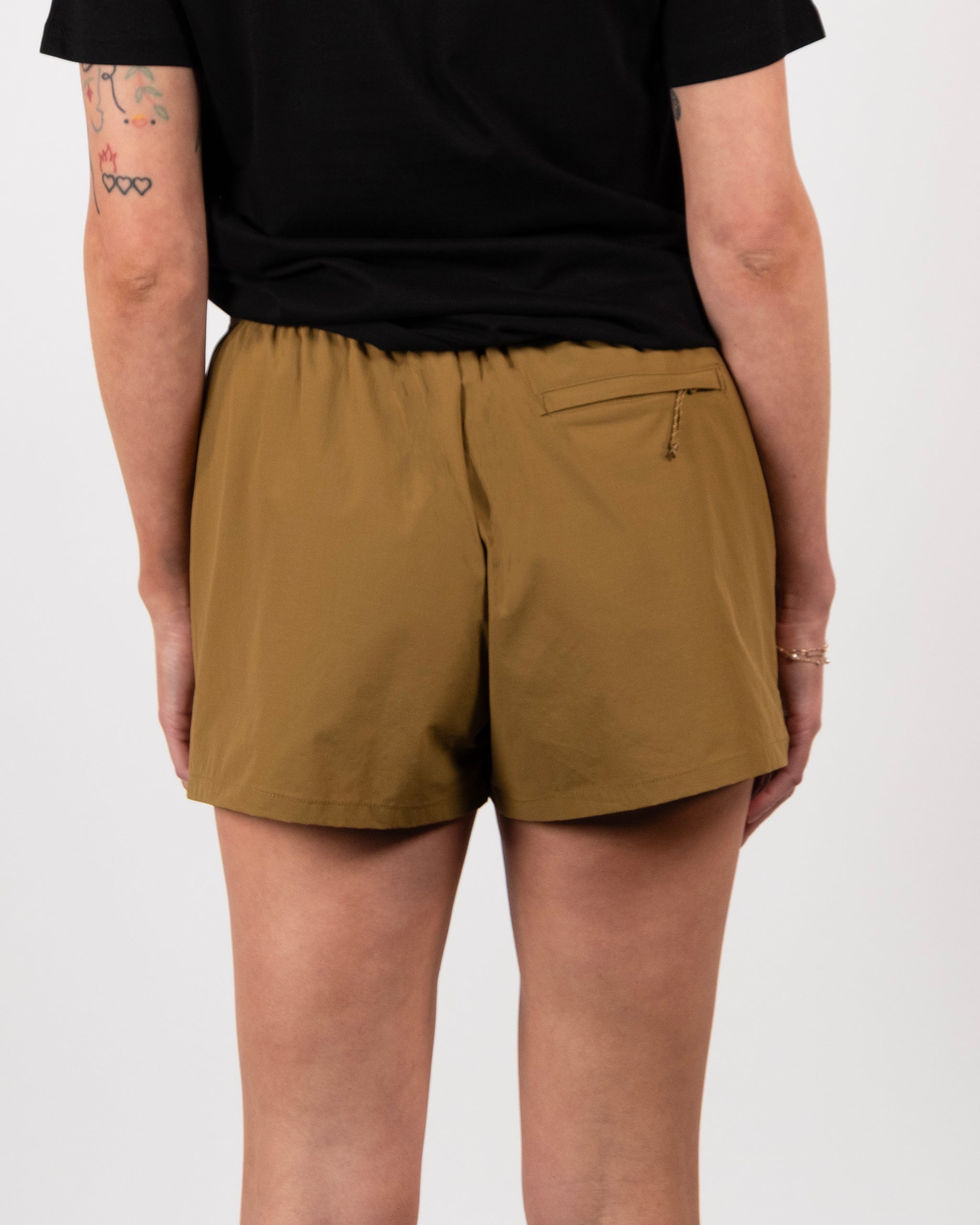 A person wearing a black shirt and BEARDEDGOAT Summit Short is shown from the back. The lightweight shorts have an elastic waistband, durable ripstop fabric, and a right zippered pocket. Tattoos can be seen on their left arm.
