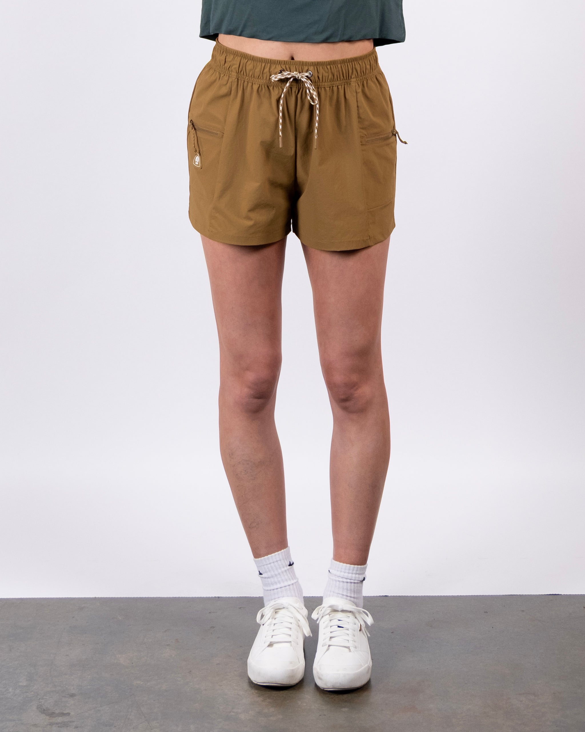 A person models the tan BEARDEDGOAT Summit Short, crafted from lightweight ripstop fabric, paired with white socks and sneakers against a plain white background. Their upper body is partially out of the frame.