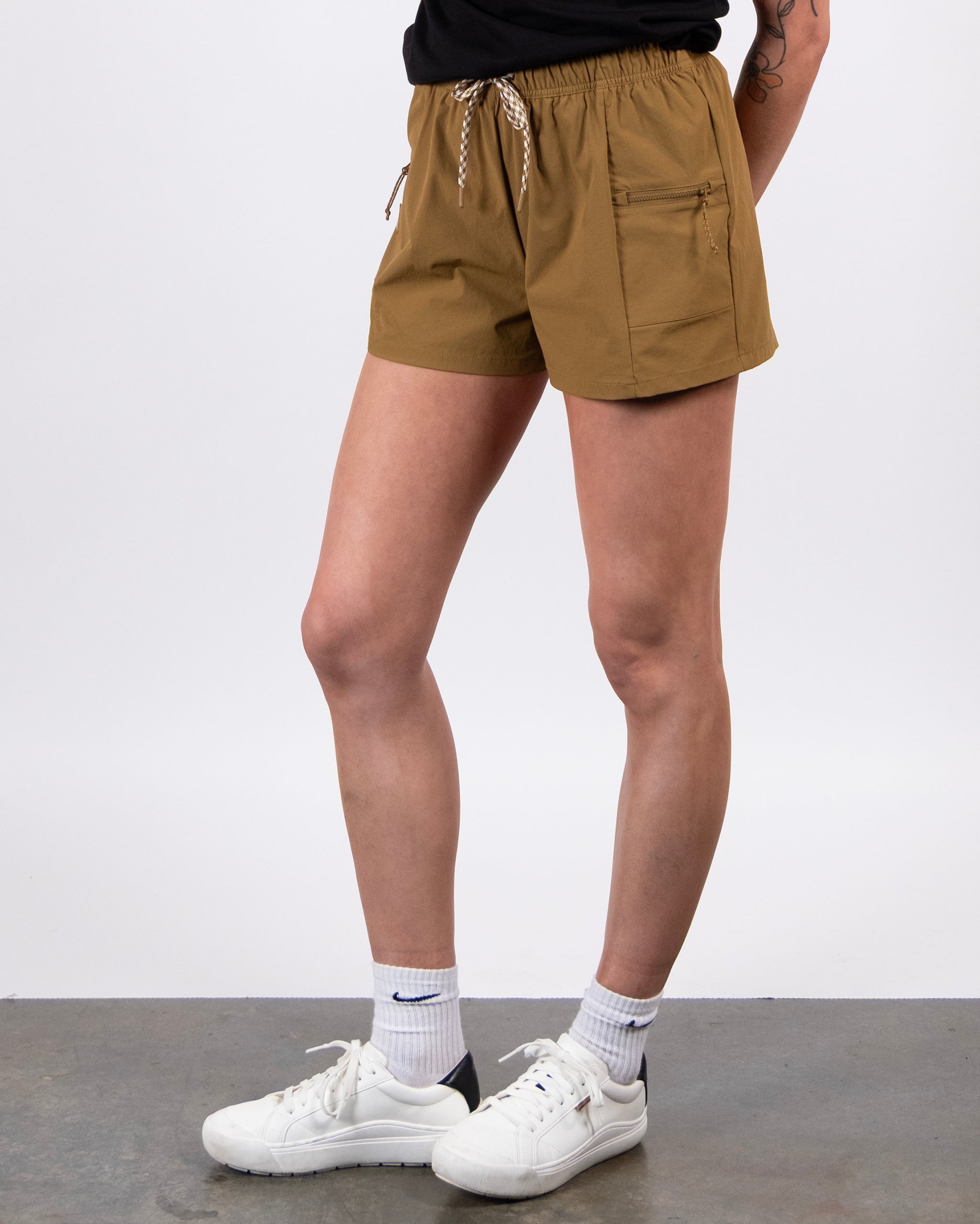 A person stands on a gray floor wearing BEARDEDGOAT Summit Short lightweight ripstop shorts in light brown, a black shirt, white socks, and white sneakers. The setting is plain white, with a tattooed arm partially visible.