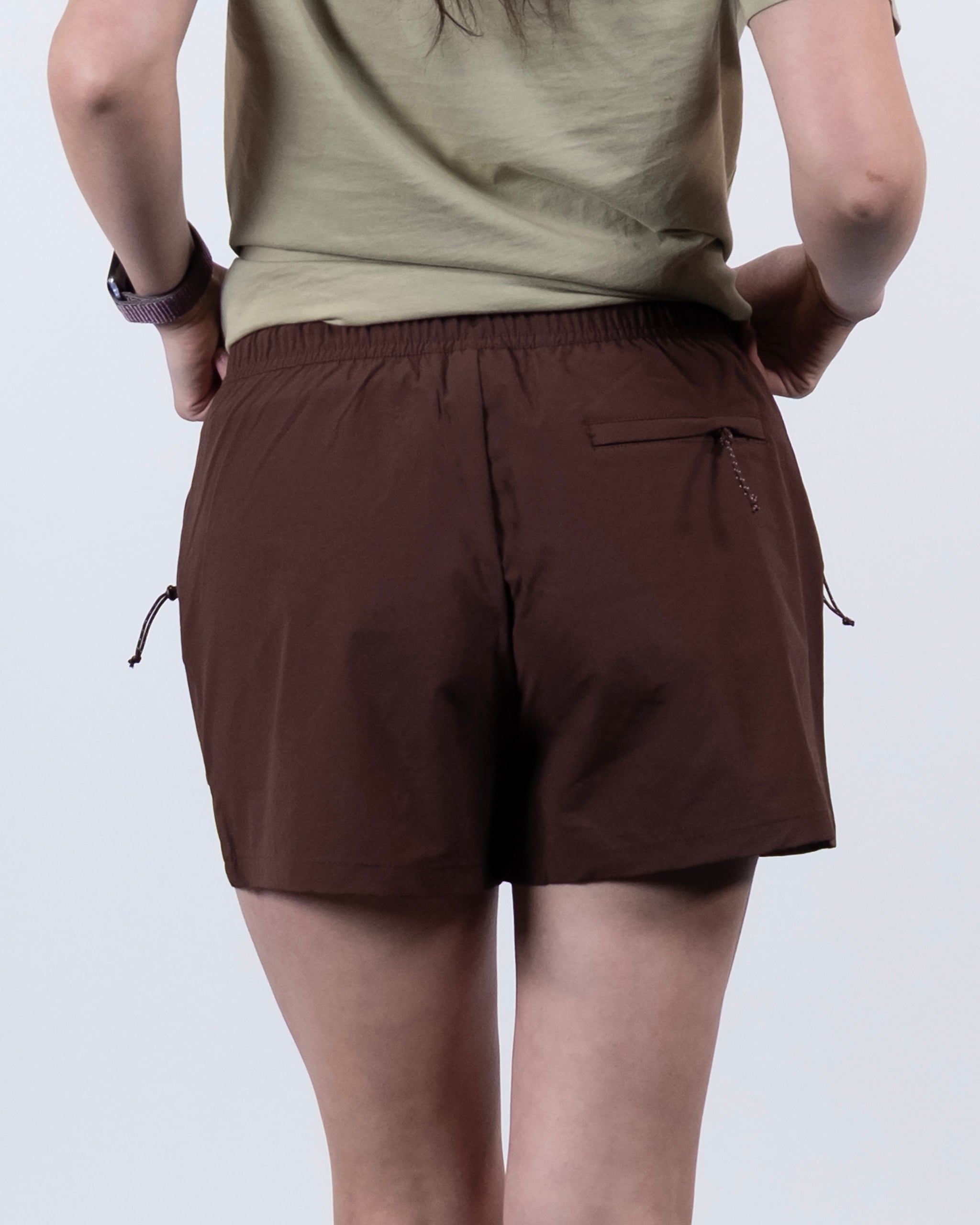 A person stands with their back to the camera, wearing a green t-shirt and BEARDEDGOAT Summit Short—lightweight ripstop shorts with side drawstrings and a zippered back pocket.