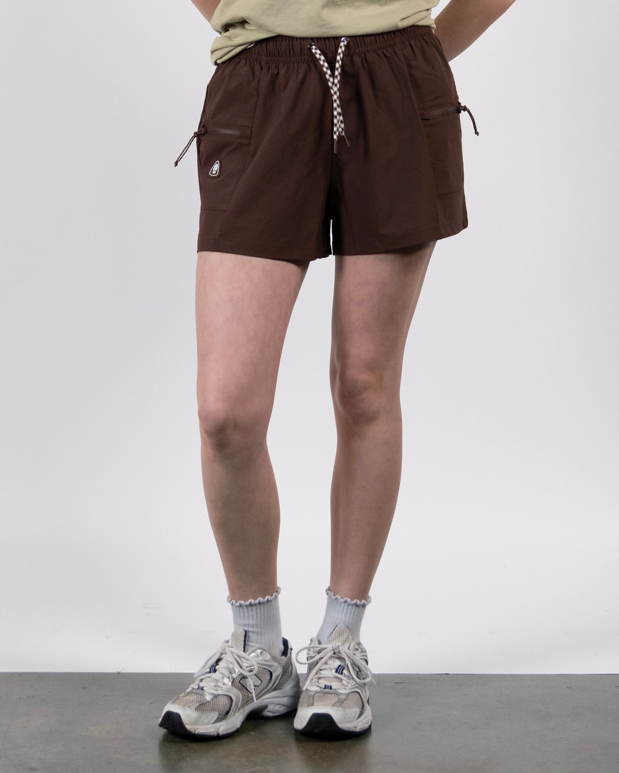 A person wearing BEARDEDGOAT Summit Short trail shorts made from lightweight ripstop fabric, light gray frilled socks, gray sneakers, and a tan shirt stands against a plain white background.
