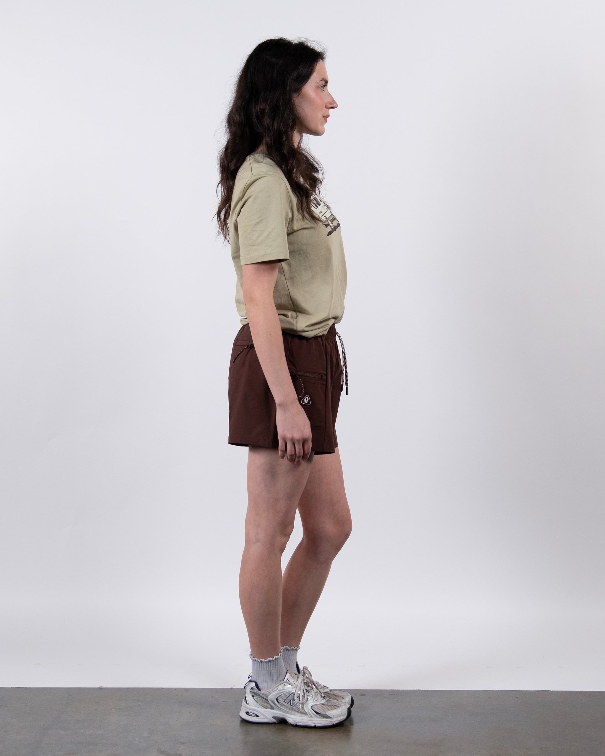 A woman in profile against a plain white background wears a beige t-shirt, BEARDEDGOAT Summit Short made from durable ripstop fabric, plus white socks and sneakers.