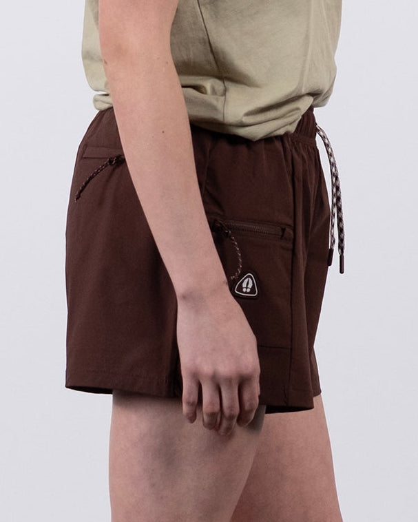 A woman stands in profile against a plain white background, wearing a beige t-shirt, BEARDEDGOAT Summit Short made from lightweight ripstop fabric in brown, along with gray socks and white sneakers.