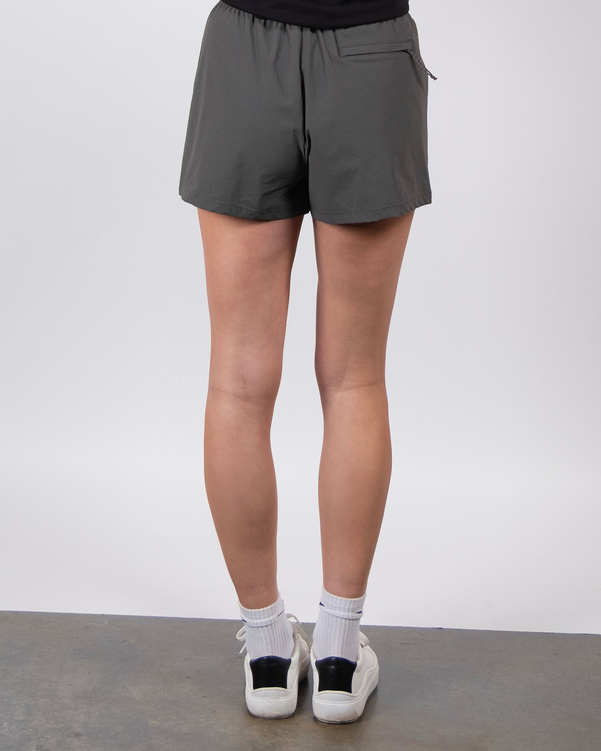 A person faces away, wearing BEARDEDGOAT Summit Short in dark gray ripstop fabric with white socks and sneakers, standing on a gray floor against a plain light background.