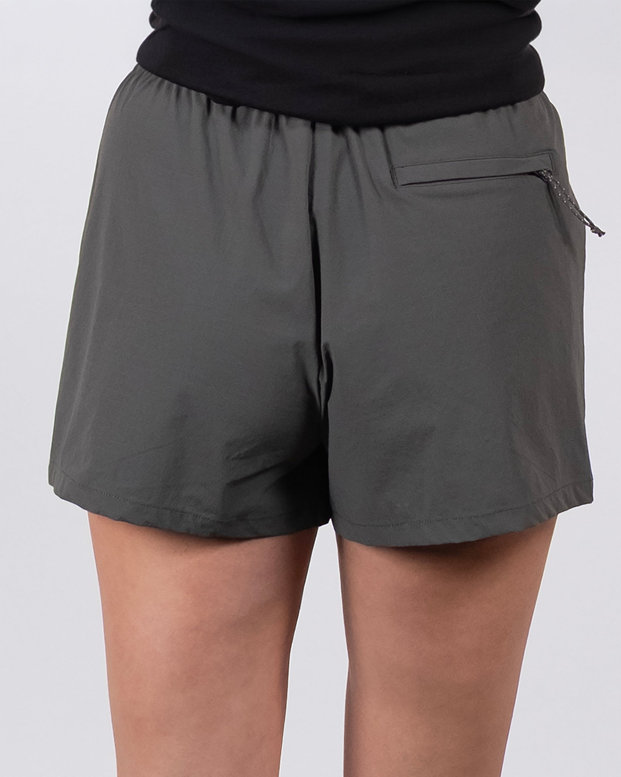 A person wears BEARDEDGOAT Summit Short trail shorts in dark gray, featuring an elastic waistband, small right-side zippered pocket, and lightweight ripstop fabric, standing against a plain light background.