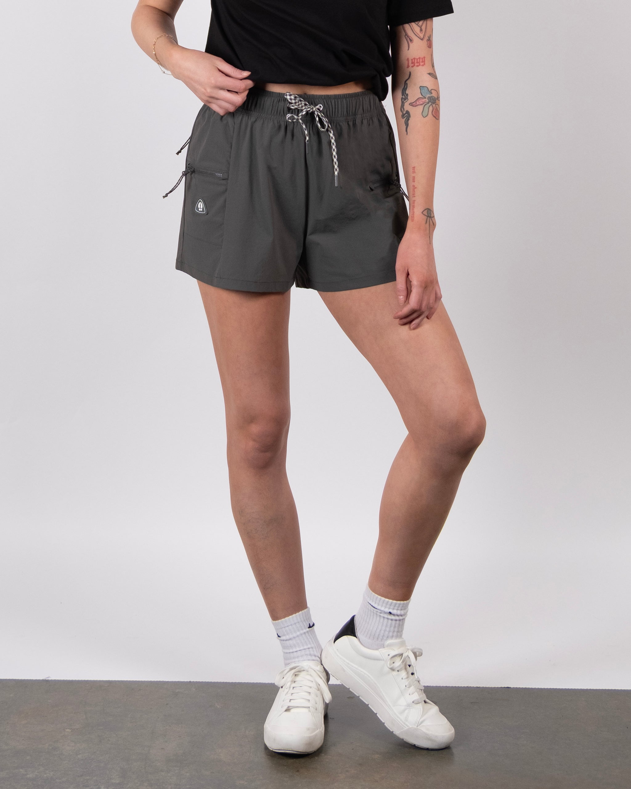 A person wearing a black t-shirt, BEARDEDGOAT Summit Short in gray lightweight ripstop with drawstring, white socks, and white sneakers stands against a plain background. Tattoos are visible on their left arm and right thigh.