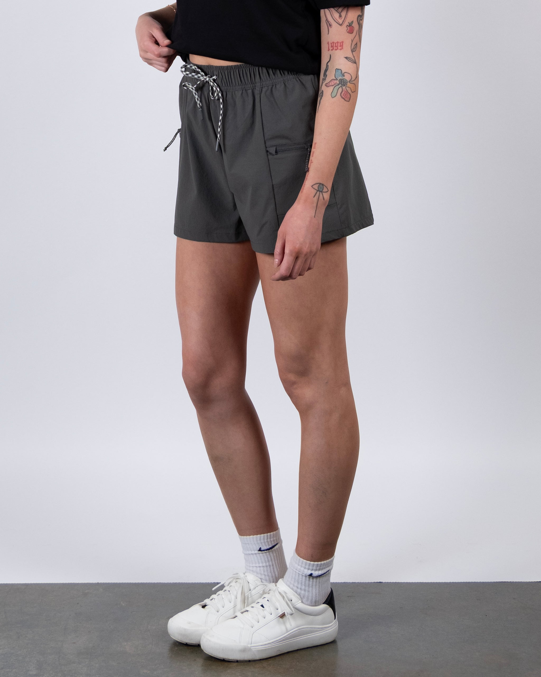 A person in a black t-shirt, BEARDEDGOAT Summit Short in gray ripstop fabric, white socks, and white sneakers stands against a plain background, showing tattooed arms.