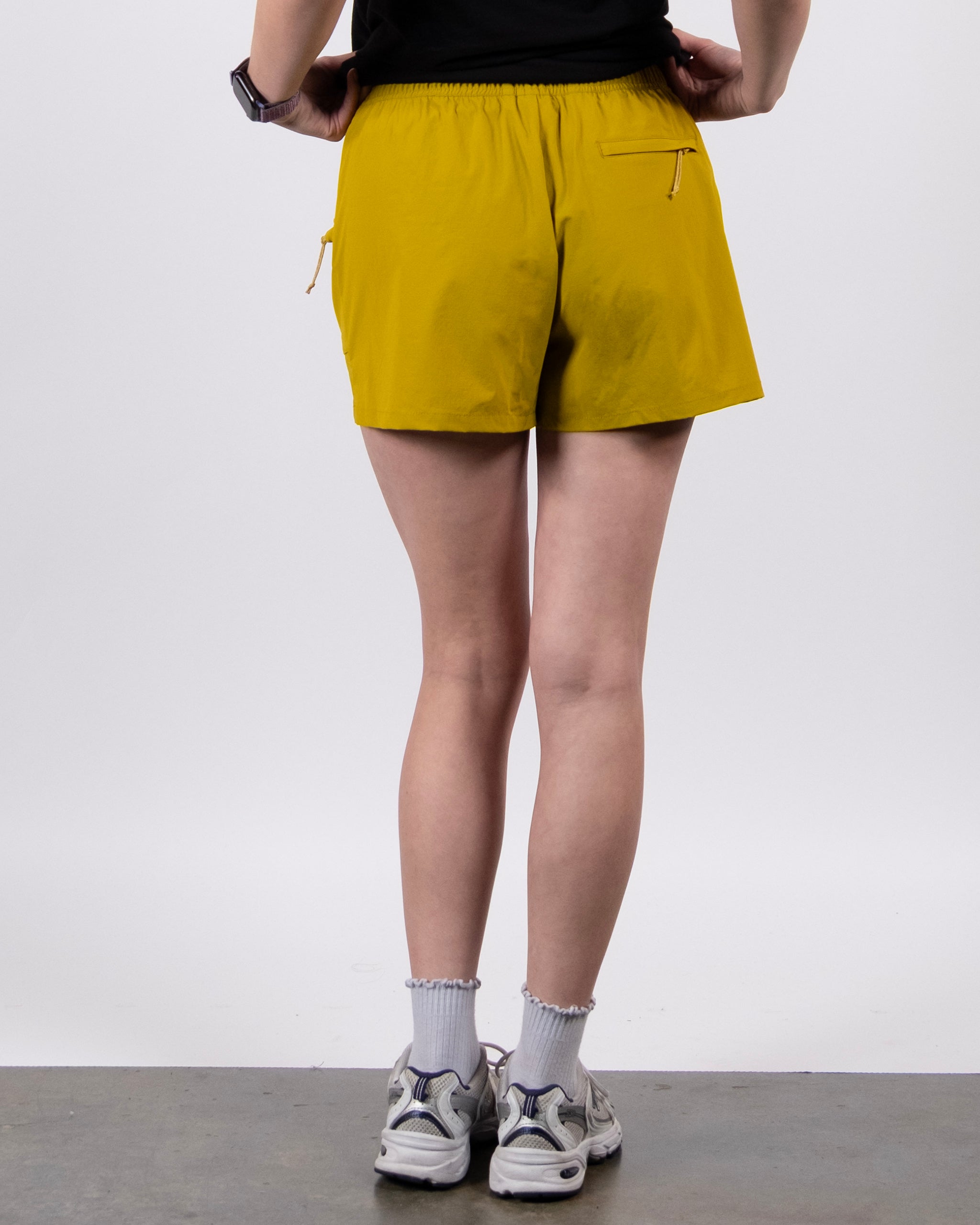 A person faces away, wearing BEARDEDGOAT’s yellow Summit Short in ripstop fabric with zippered back pockets, paired with a black top, ruffled white socks, and white sneakers. The background is plain and light.