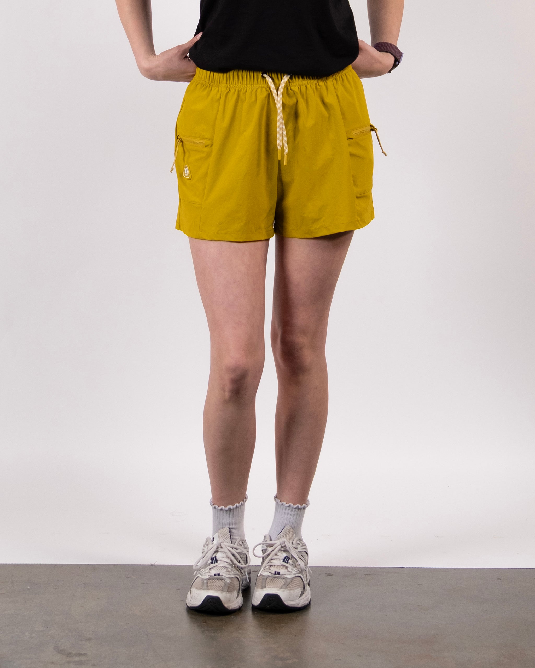 A person stands with hands on hips, wearing a black top and BEARDEDGOAT Summit Short in Golden Palm made from ripstop fabric, featuring side pockets, plus white socks and sneakers against a plain background.