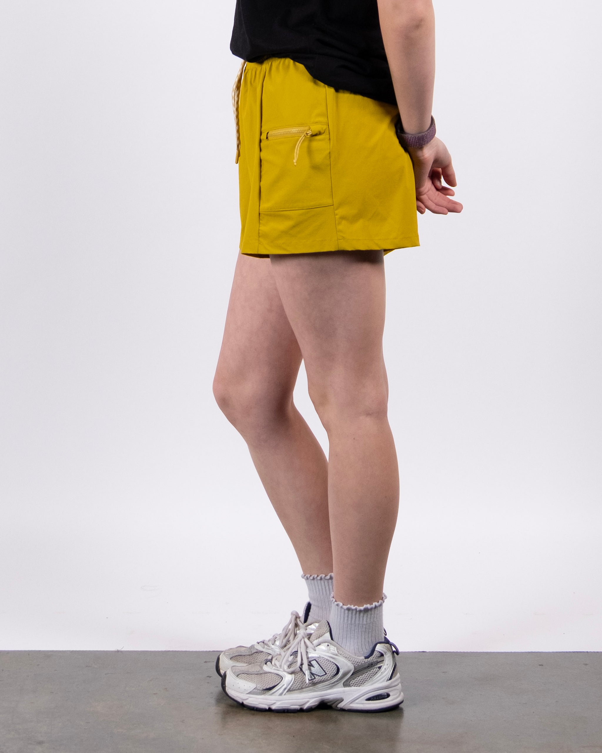 A person stands sideways against a plain background, wearing a black shirt, BEARDEDGOAT Summit Shorts made from ripstop fabric with front pockets, white socks, and white sneakers. Their arms are relaxed behind their back.