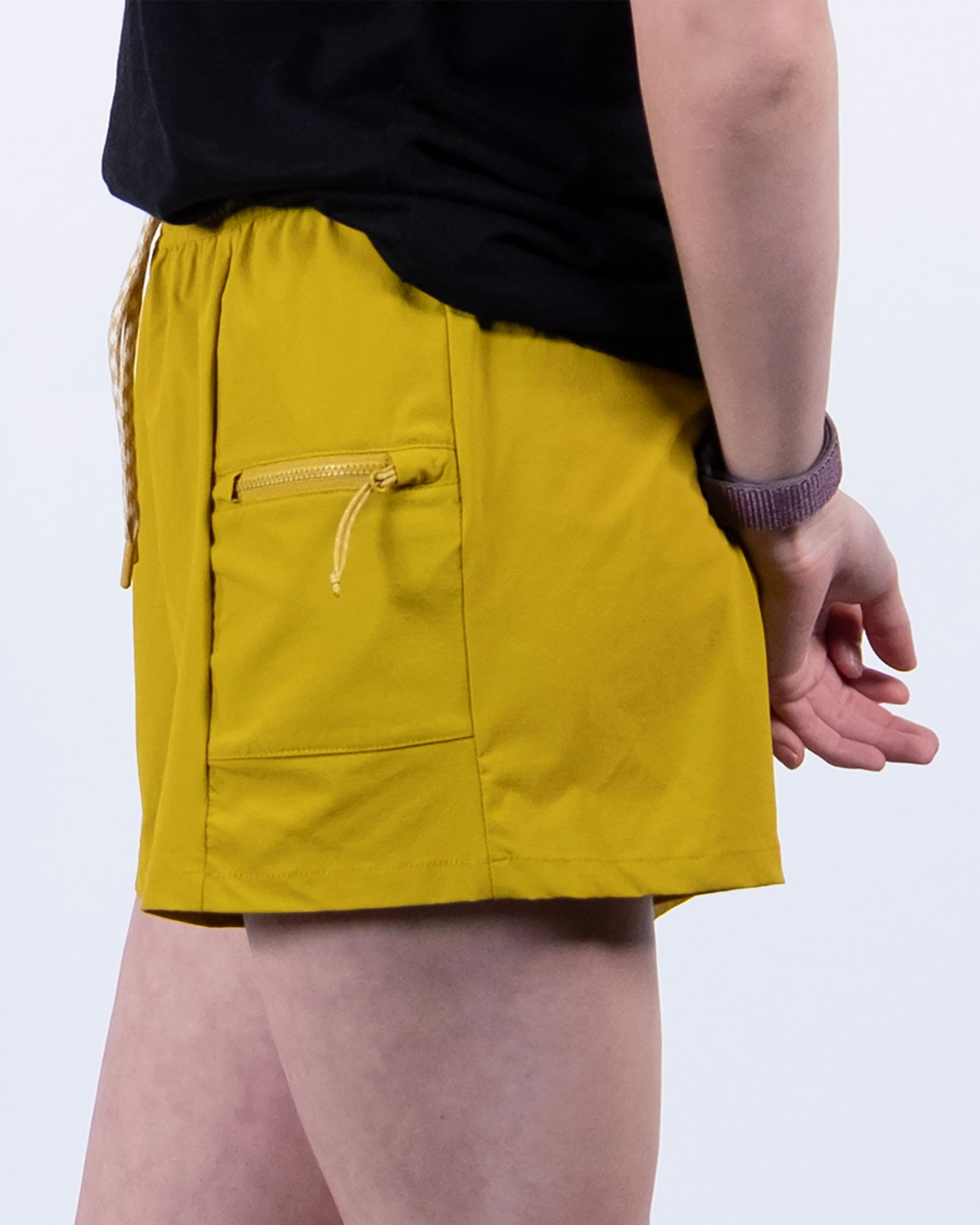 A person in BEARDEDGOAT Summit Short trail shorts in mustard yellow ripstop fabric, a black top, and a purple watch stands against a plain white background.