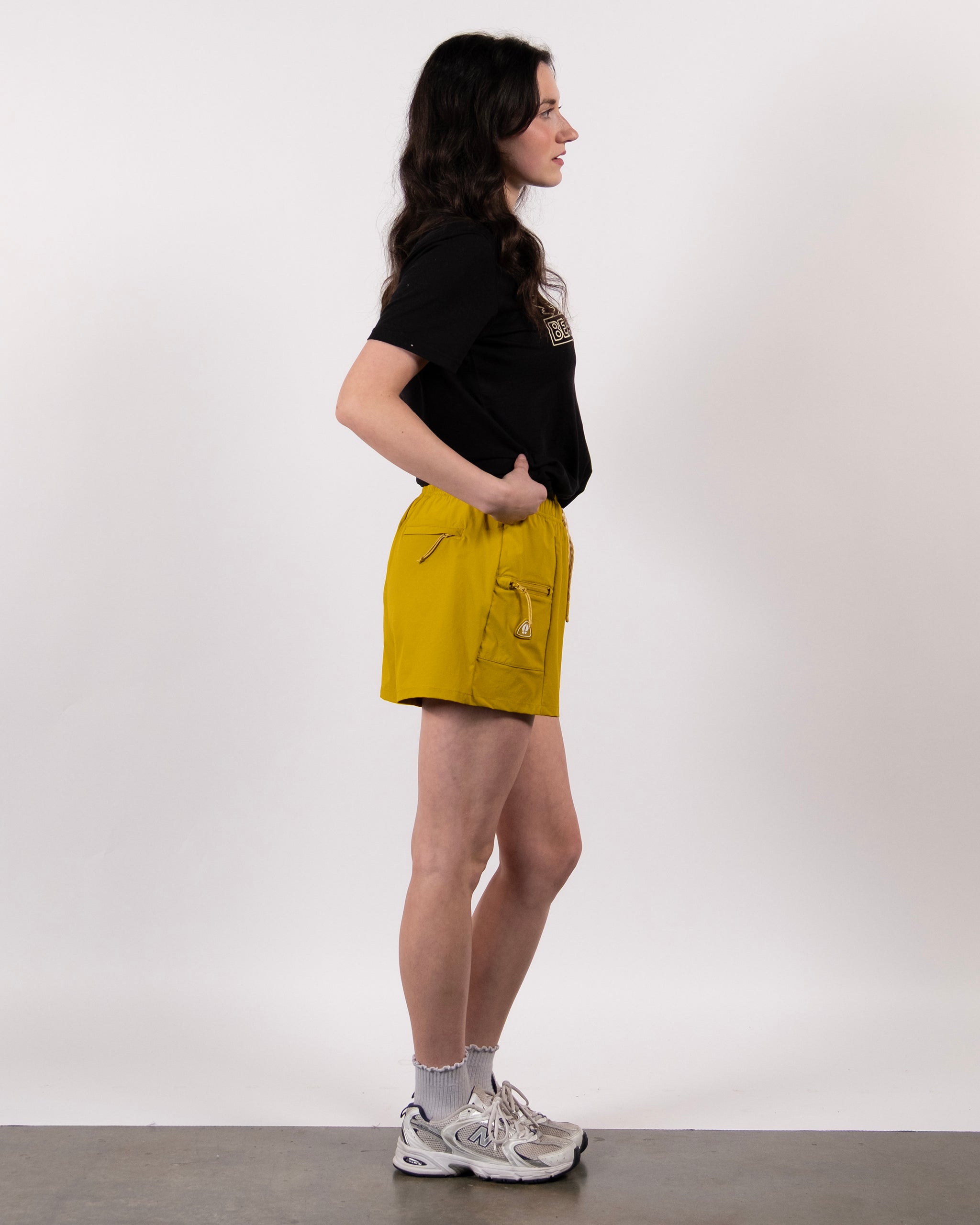 A person with long, wavy dark hair stands in profile against a white background, wearing a black t-shirt and bright yellow BEARDEDGOAT Summit Short made from lightweight ripstop fabric, with gray socks and white sneakers.