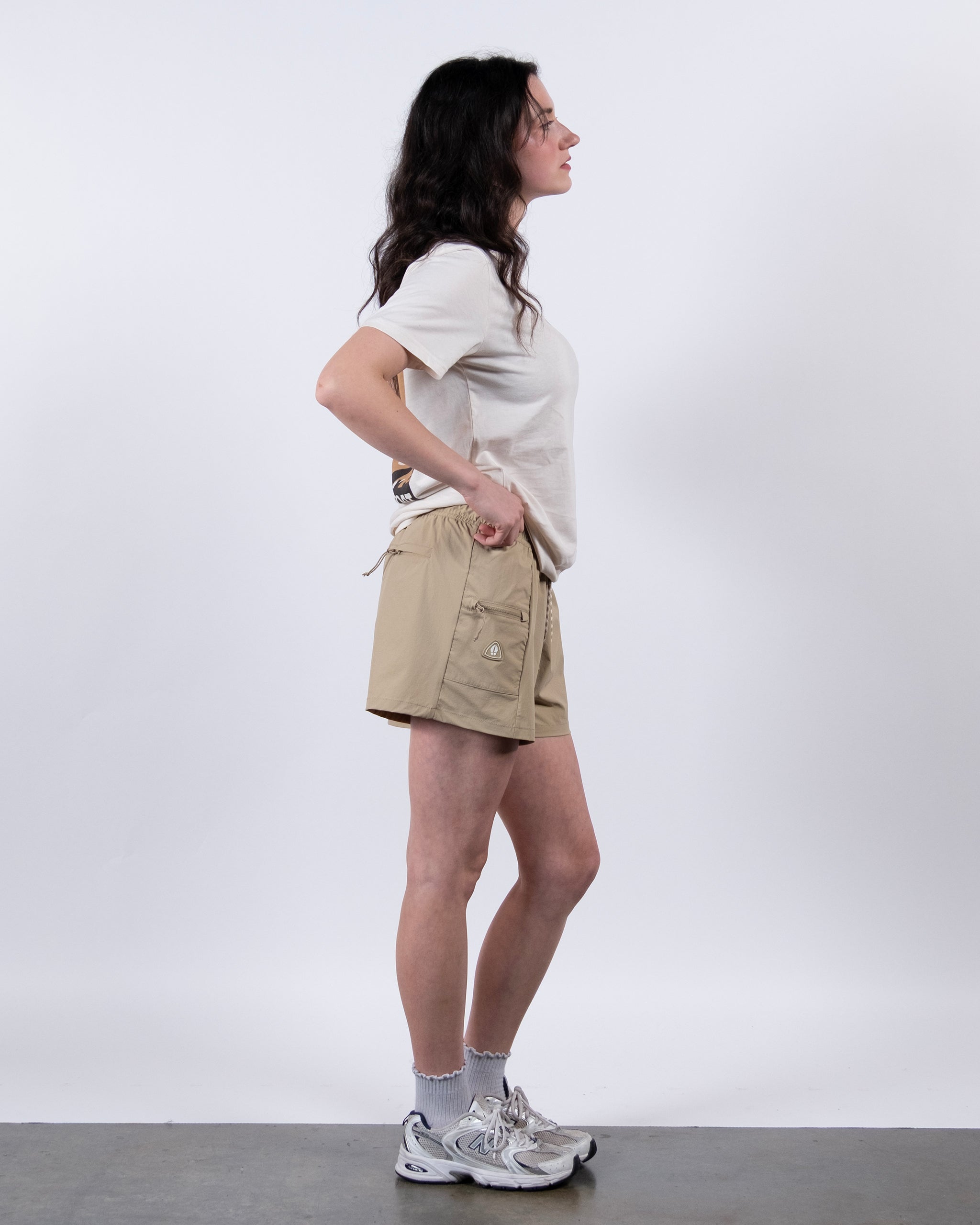 A woman stands in profile against a white background, wearing a beige t-shirt and BEARDEDGOAT Summit Short—lightweight, pocketed shorts crafted from durable ripstop fabric. She pairs the look with white socks, athletic shoes, and loose hair.