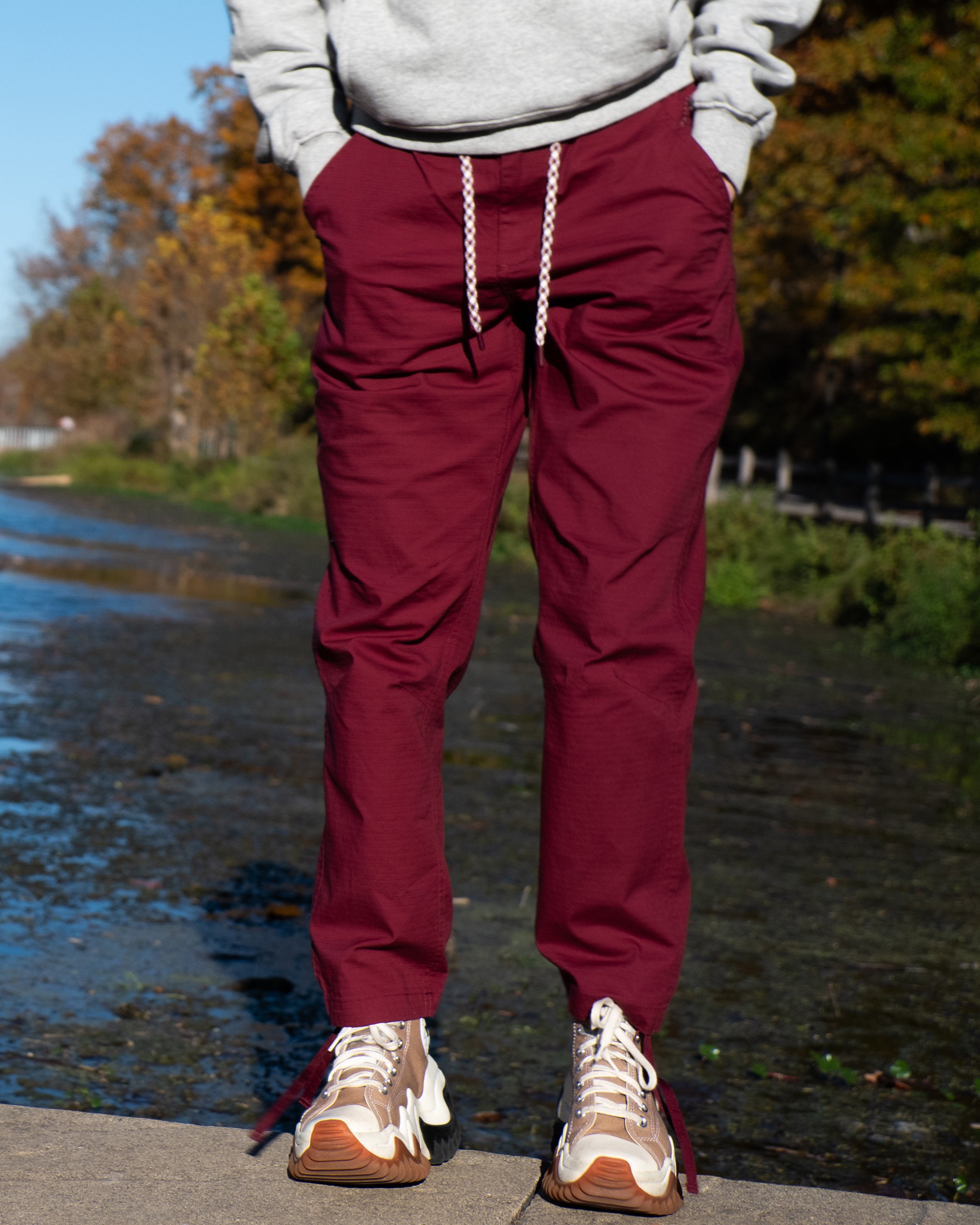 A person stands outdoors near water in BEARDEDGOAT Women's Canopy™ Pant, made of stretch cotton ripstop, paired with tan and white sneakers and a light gray sweatshirt. Their hands are in their pockets; autumn trees create a scenic backdrop.