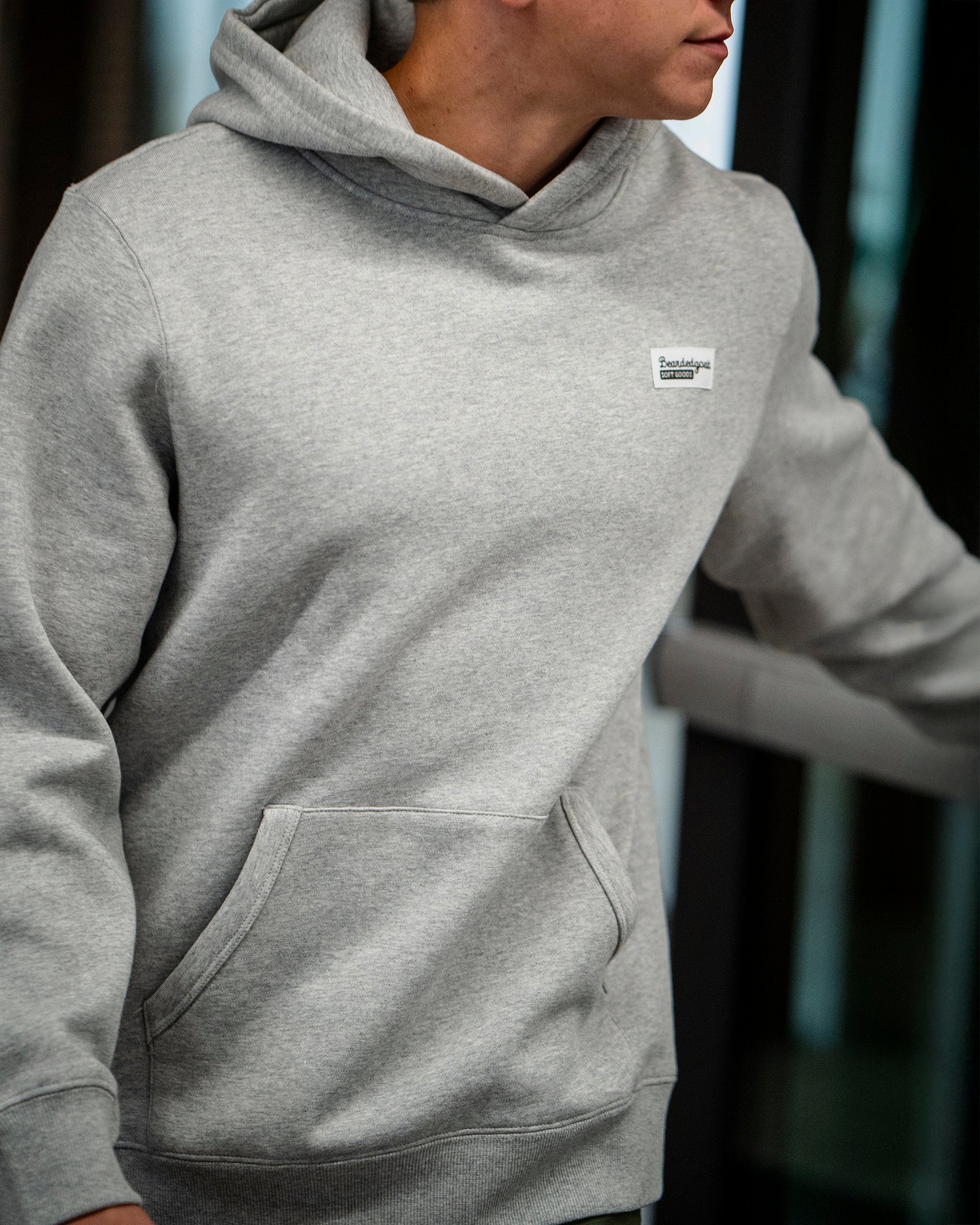A person wears The VOID™ Hoodie by beardedgoat in light gray, made from premium heavyweight cotton fleece. The hoodie has a front pocket and a small chest patch; they stand indoors, facing partially to the side with their face mostly out of frame.
