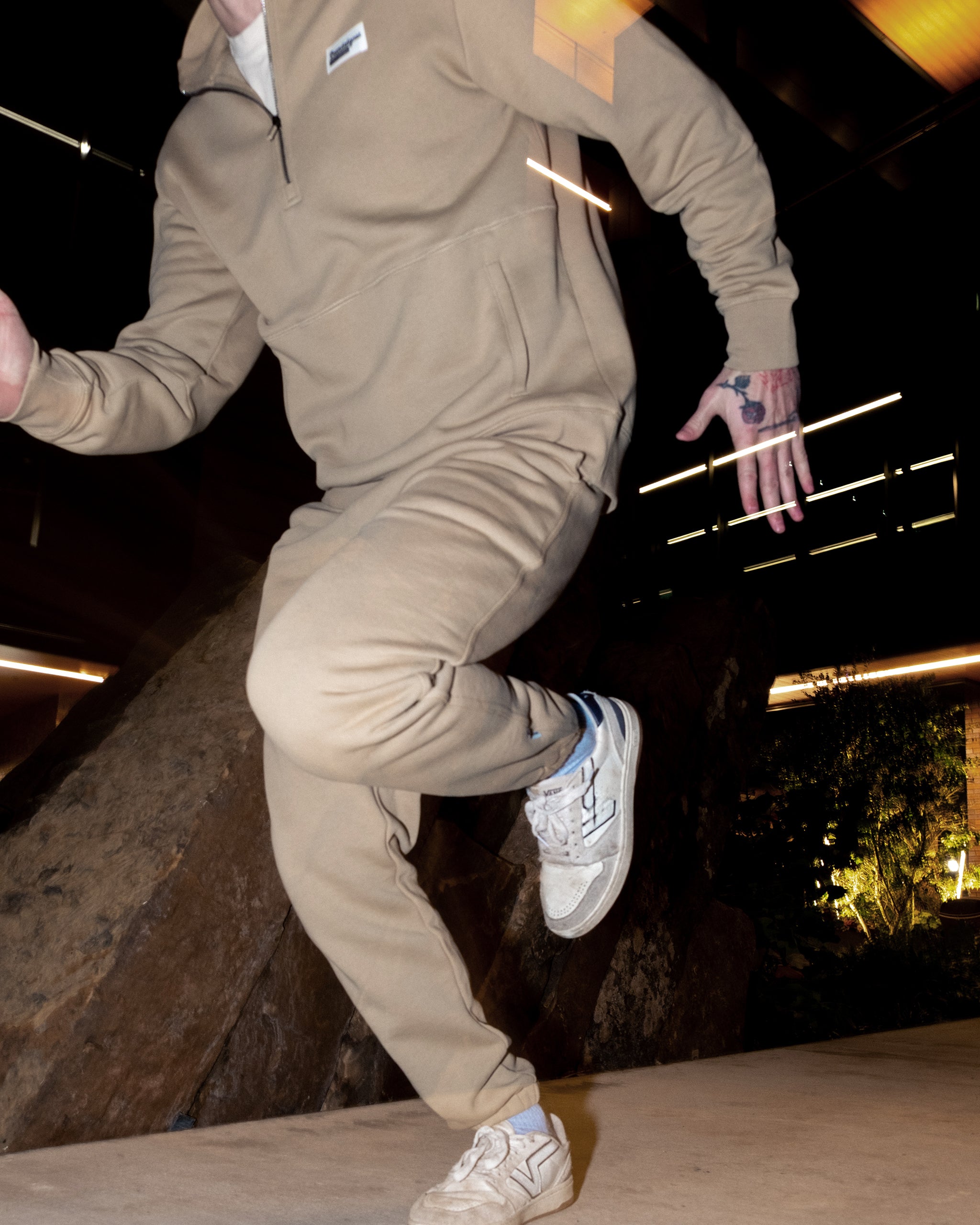 Someone wearing beige beardedgoat VOID™ Sweatpants and white sneakers is captured mid-motion indoors, with blurred lights and a large rock in the background. Their face remains unseen.