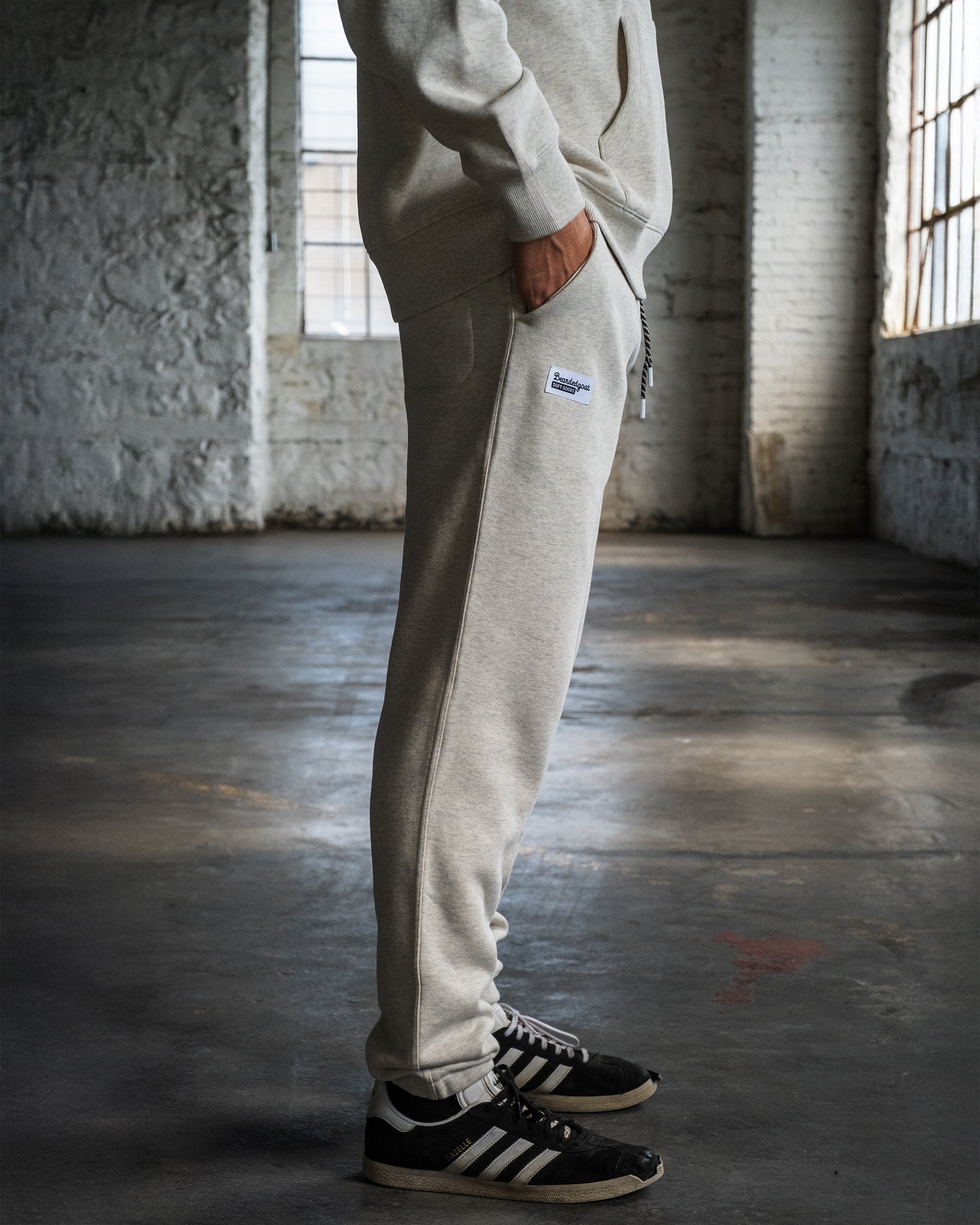 A person in beardedgoat's VOID™ Sweatpant and black Adidas sneakers stands in a sunlit, industrial-style room with textured white brick walls and large windows.