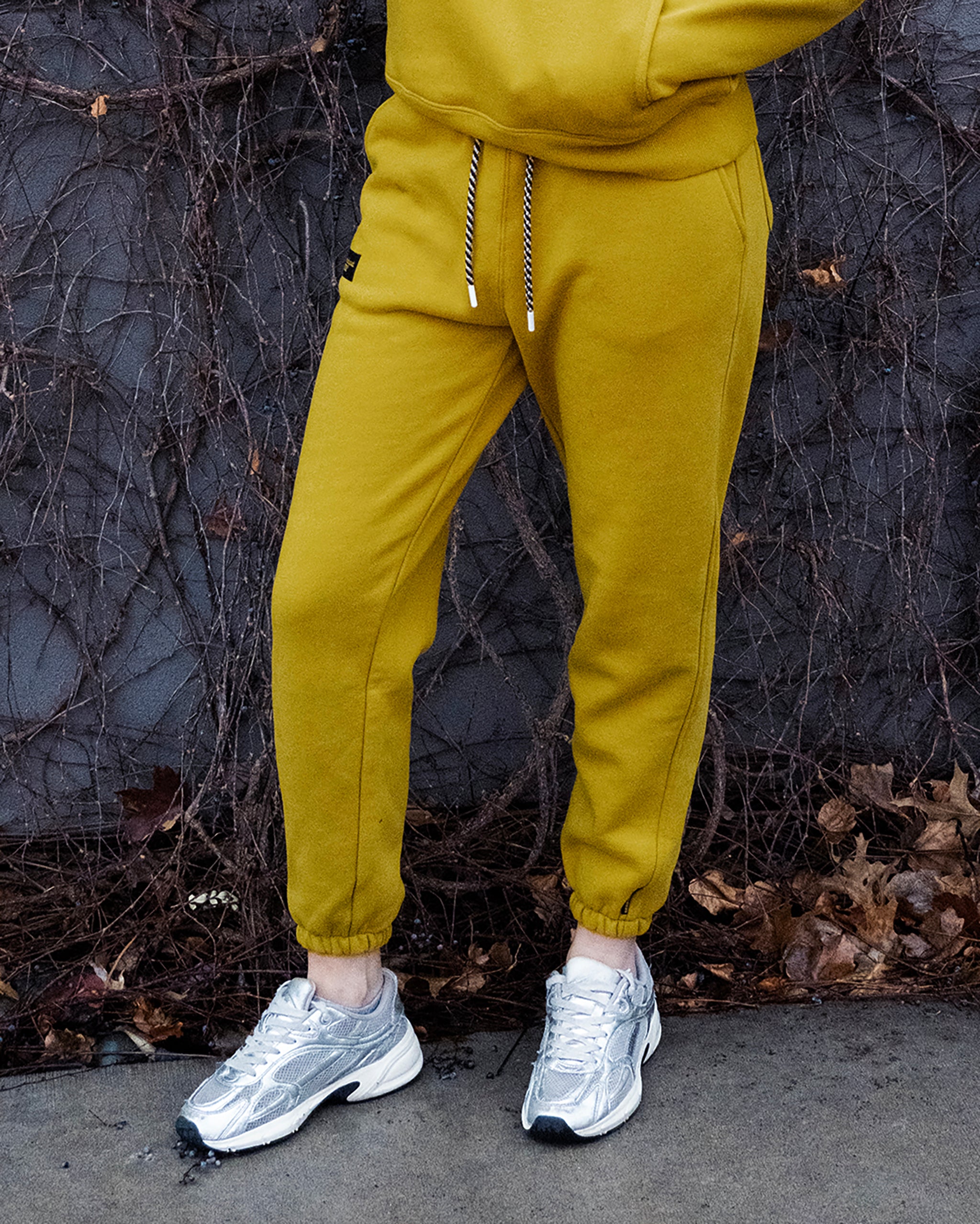 Wearing BEARDEDGOAT’s NEW — Overnight™ Classic Jogger in mustard yellow with a high-rise waistband and silver sneakers, a person stands on dry leaves before bare, tangled vines. The upper body and face are out of frame.