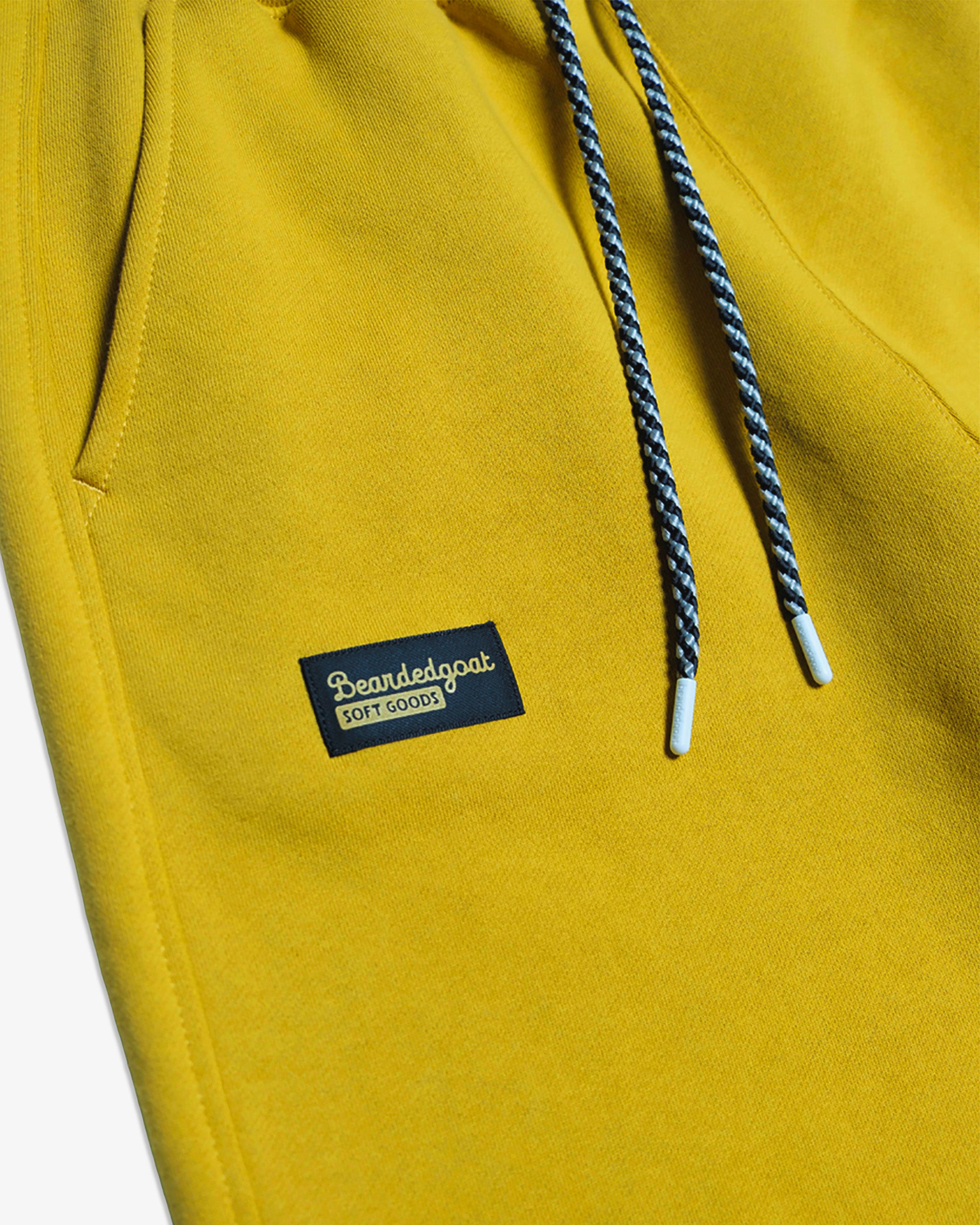 Close-up of BEARDEDGOAT NEW — Overnight™ Classic Jogger in yellow, featuring a high-rise waistband, black-and-white drawstrings, and a rectangular BEARDEDGOAT Soft Goods logo patch near the pocket.