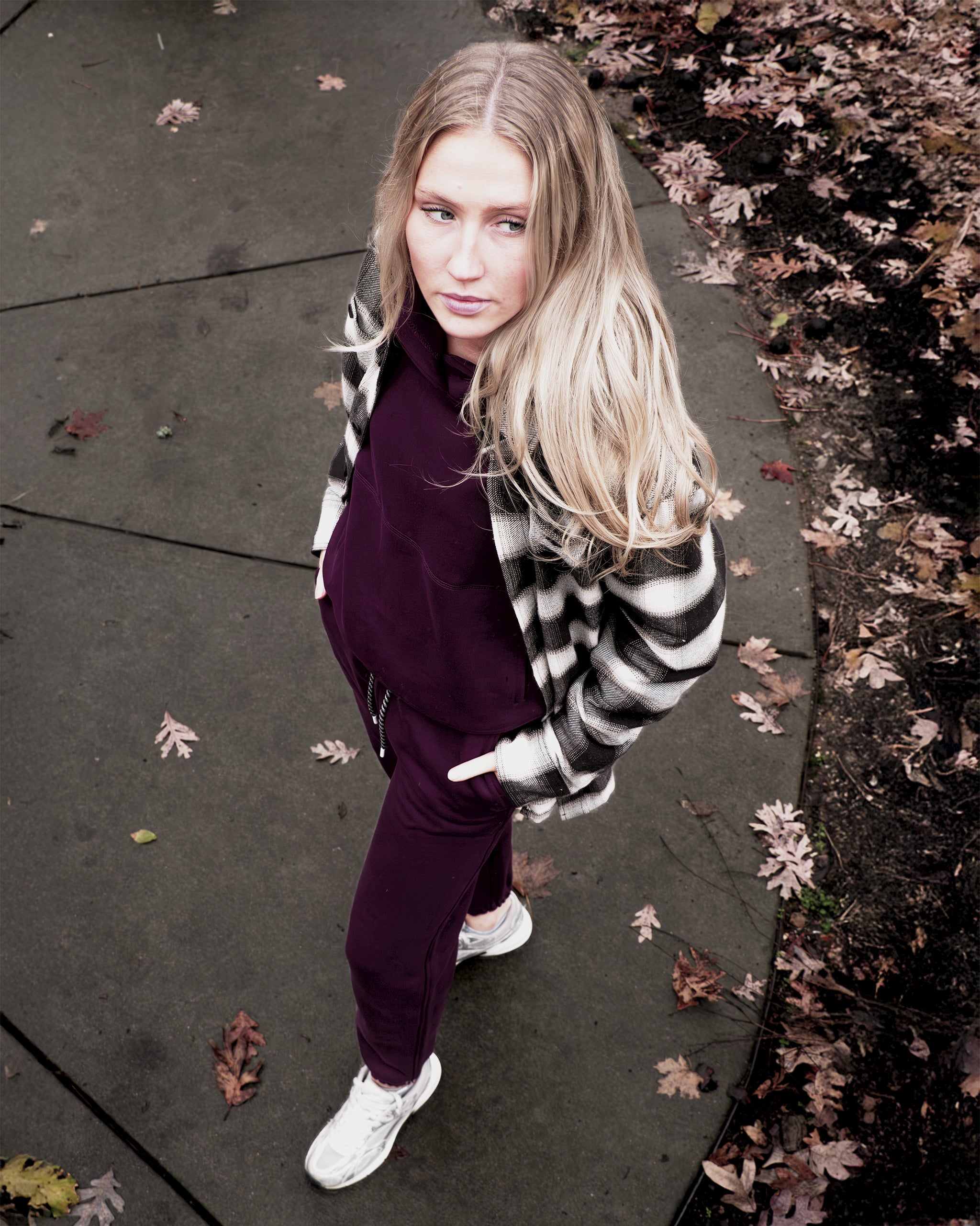 A woman with long blonde hair stands on a leaf-strewn path, wearing the beardedgoat Women's Overnight™ Hoodie, white sneakers, and a black-and-white plaid jacket, gazing up at the camera.