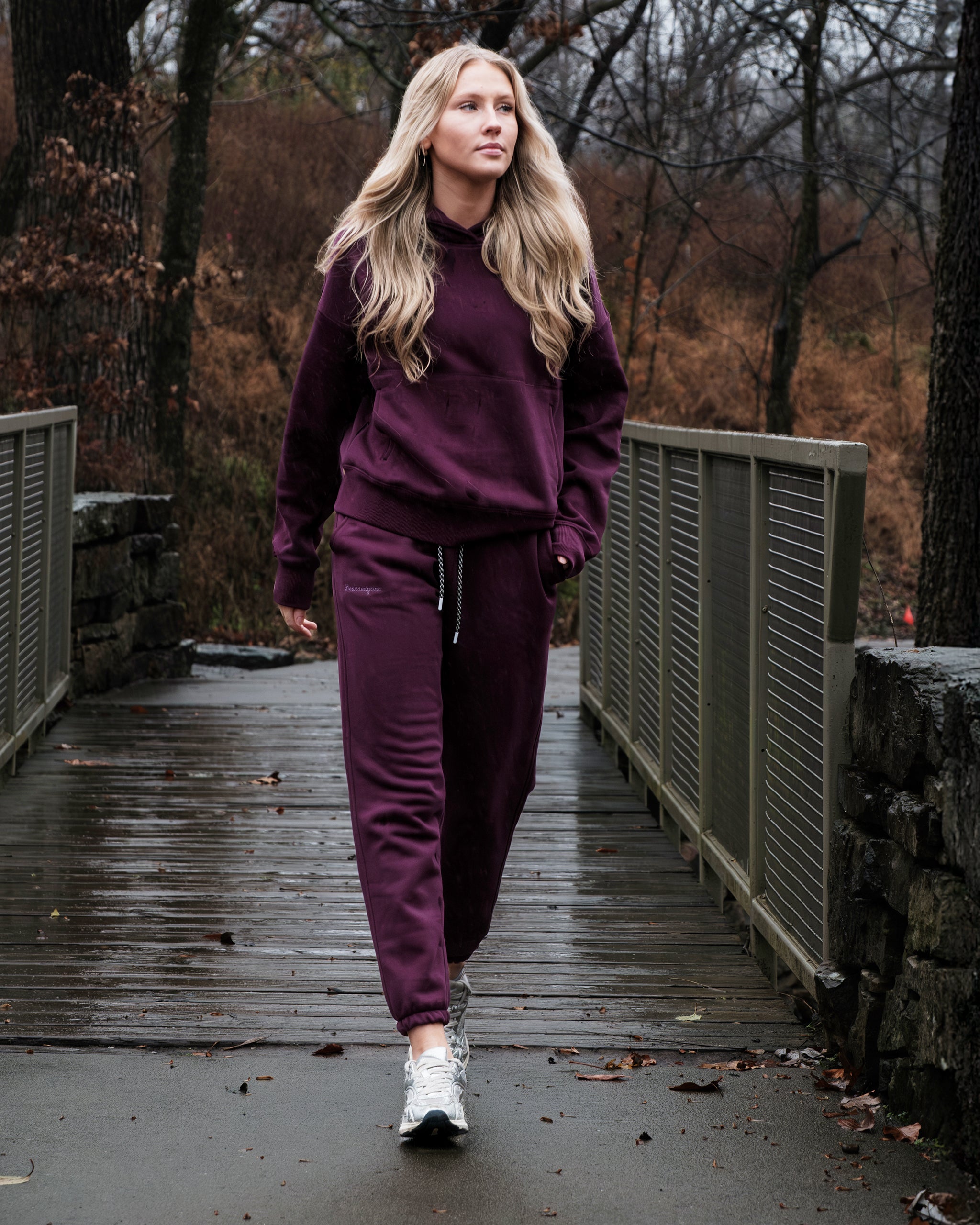 A woman with long blonde hair wears the beardedgoat Women's Overnight™ Hoodie and maroon tracksuit with white sneakers as she walks on a wet wooden bridge in a forested area on a cloudy day.