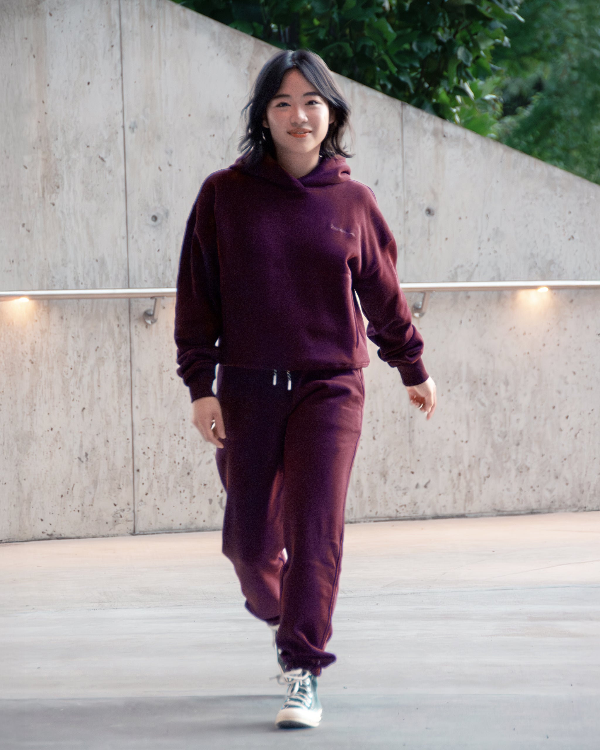 A woman with dark hair walks outdoors in front of a concrete wall with greenery, wearing BEARDEDGOAT's NEW — Overnight™ Classic Jogger and a matching burgundy sweatshirt, paired with white sneakers.